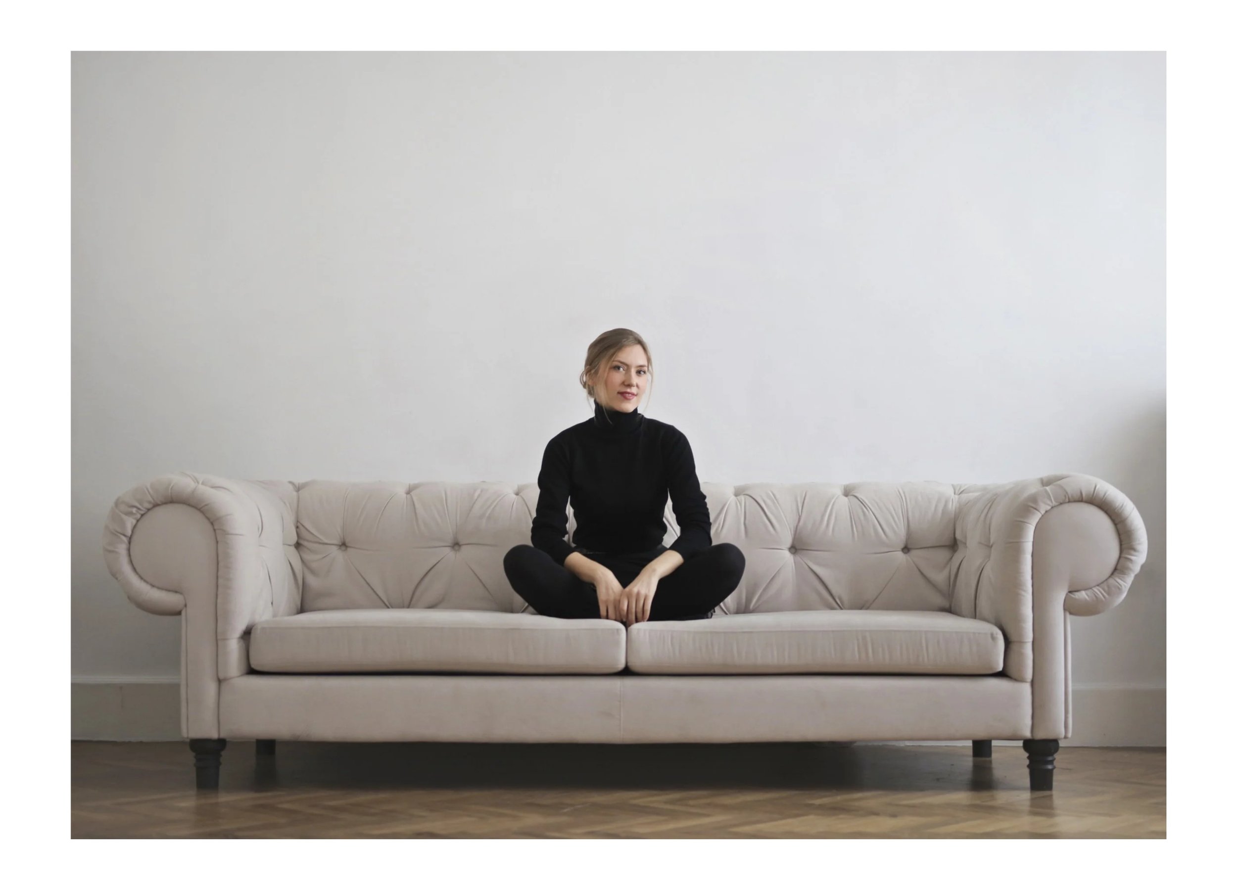 A young woman in black clothing sitting cross-legged on a beige tufted sofa in a minimalist room with a plain white wall and wooden floor.