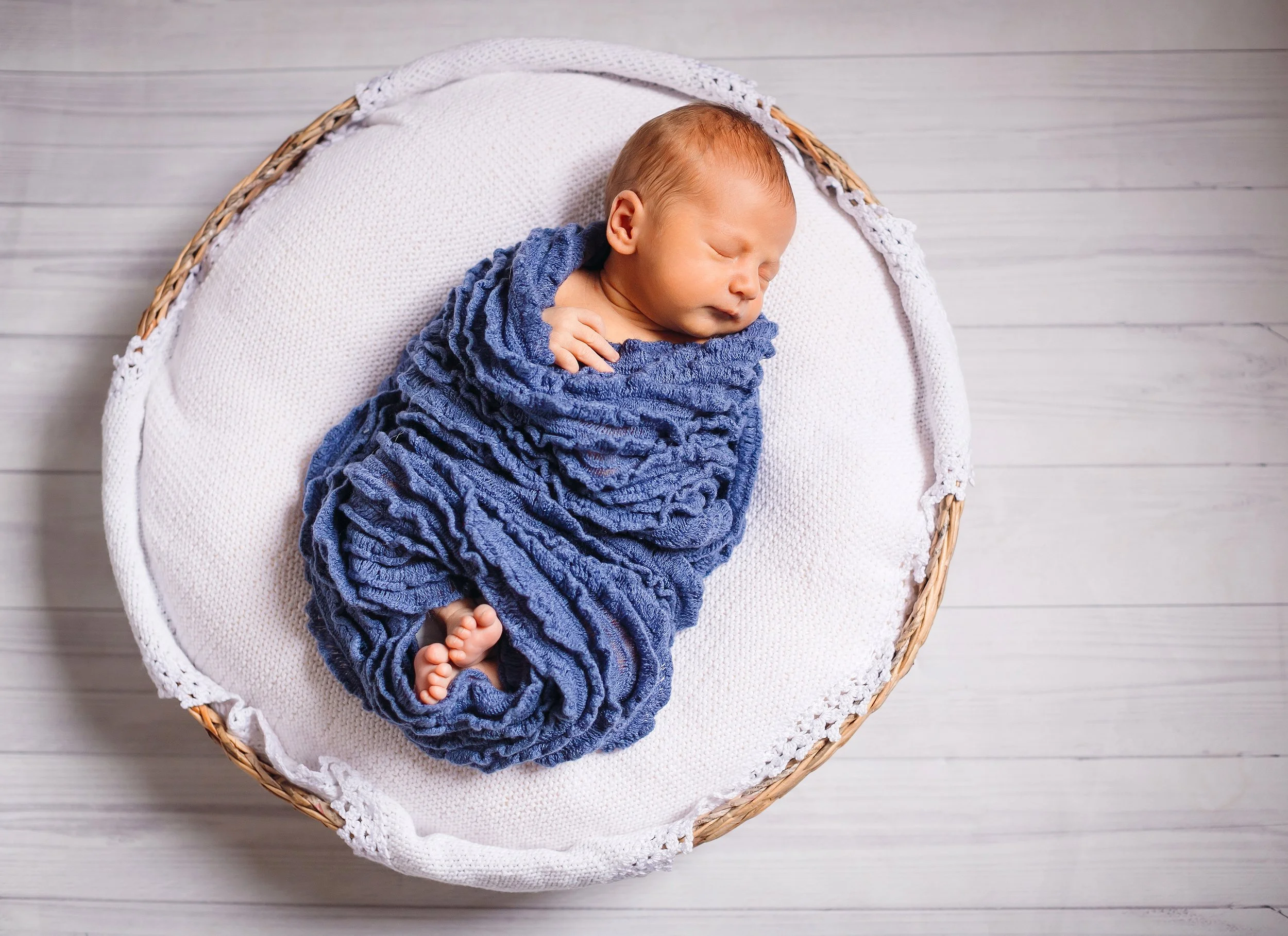 A small baby wrapped in a blue blanket, sleeping in a white basket with a lace trim, on a light wooden floor.