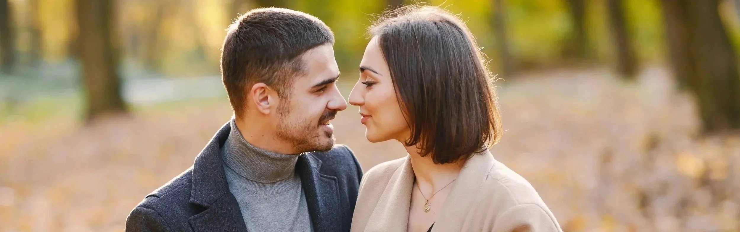 A young man and woman standing close together outdoors, facing each other with their noses touching, in a park with autumn foliage in the background.  Couples therapy page for Lady Bird Relationship Center in Austin, Texas