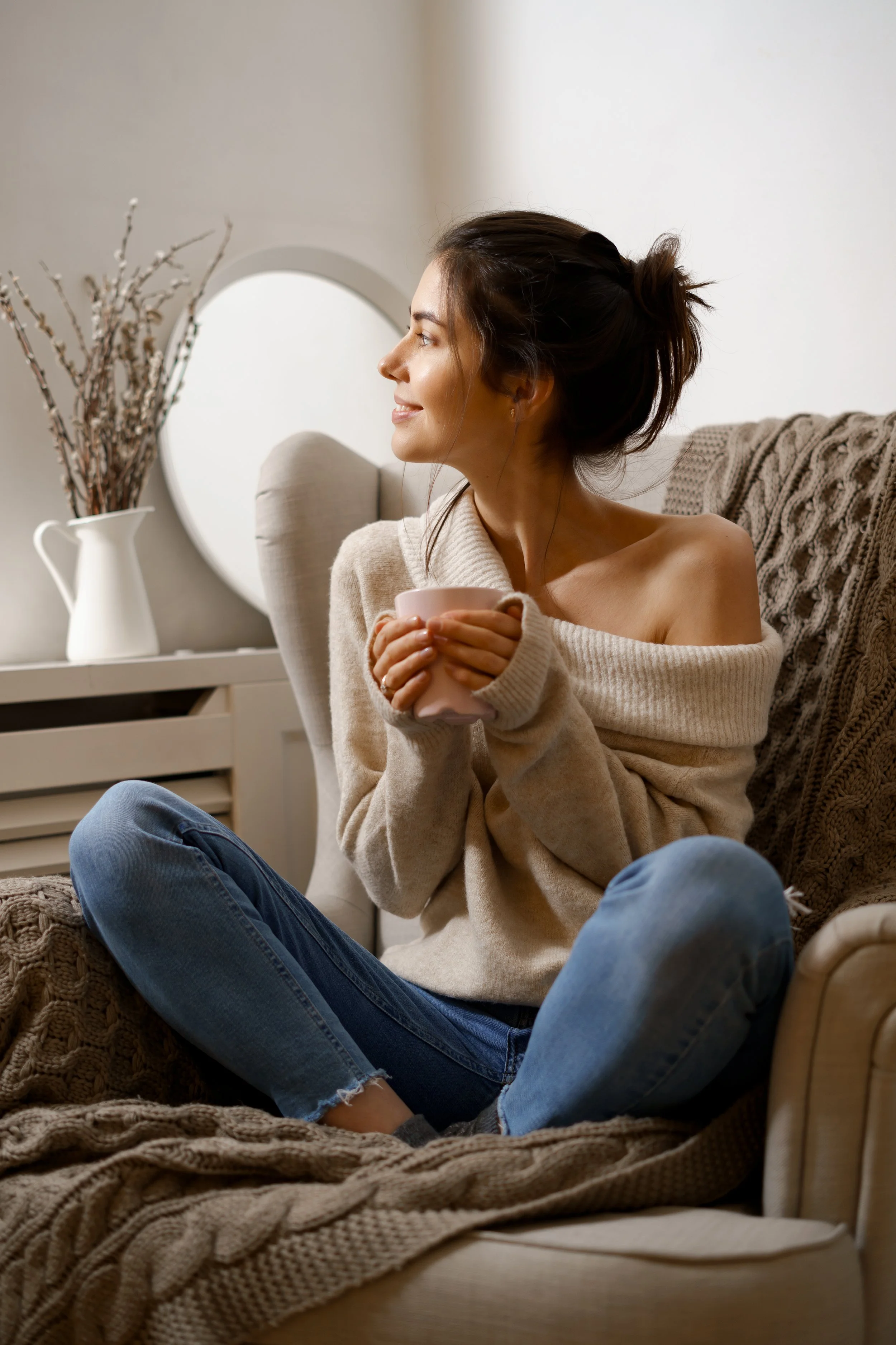 A woman with an off-shoulder beige sweater and blue jeans sitting on a cozy beige armchair, holding a pink mug, and looking to the side in a warmly lit living room.