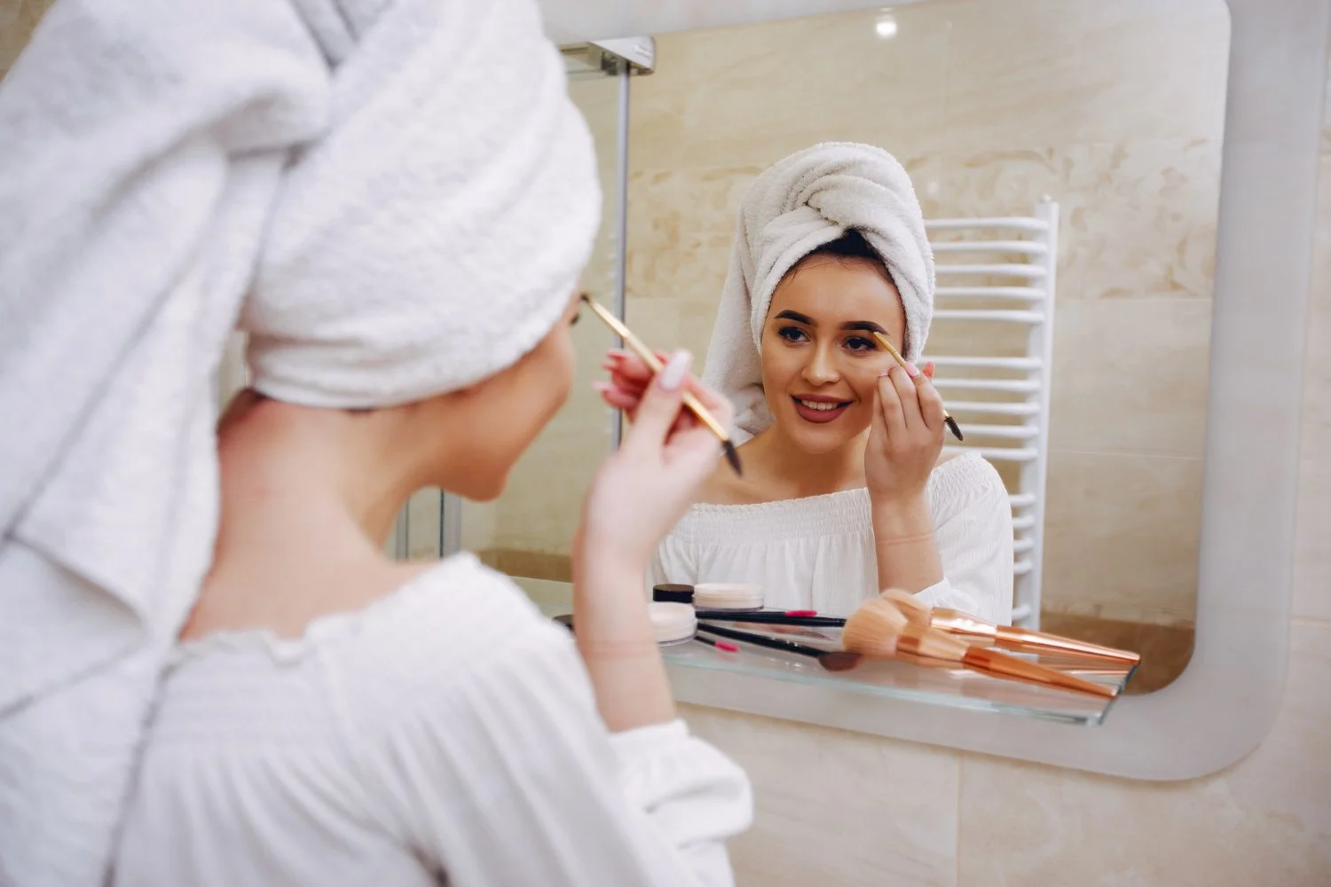 Woman in a white robe and towel on her head applying makeup in front of a mirror in a bathroom.