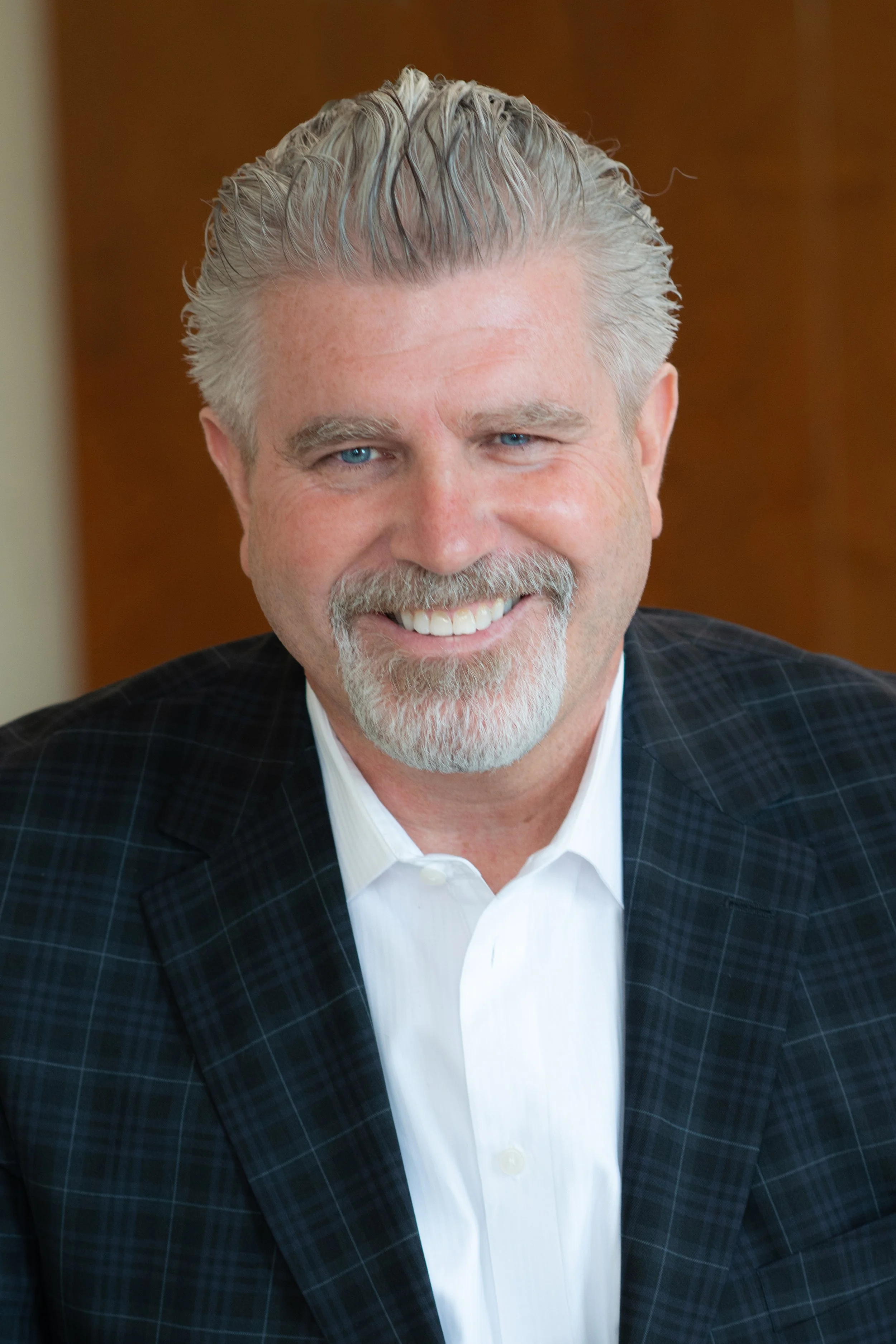 A smiling man with gray hair, blue eyes, and a beard, dressed in a white shirt and dark checked blazer, sitting indoors with a wooden background.