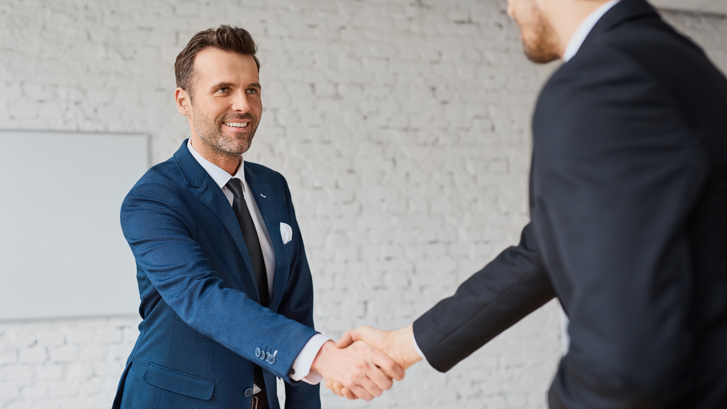 Two men in business suits shaking hands in a professional setting with a white brick wall background.
