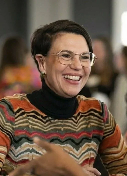 A woman with short dark hair, glasses, and a colorful striped shirt smiling in a social setting.