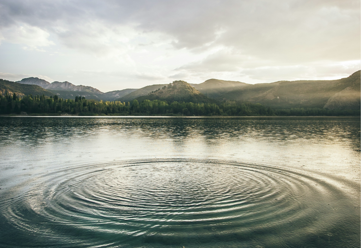 A calm lake with gentle ripples, surrounded by forested mountains under a partly cloudy sky during sunset or sunrise.