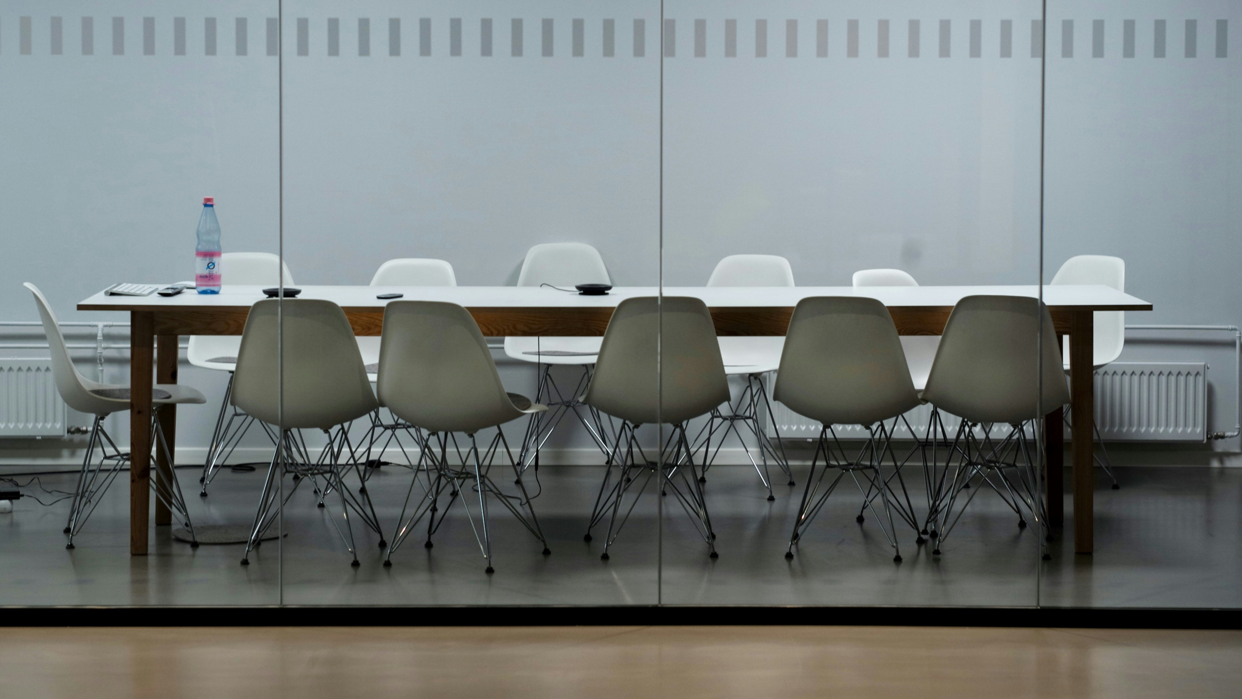 Empty conference room with a long white table, white chairs, a water bottle, paper, and electronic devices on the table, viewed through glass walls.