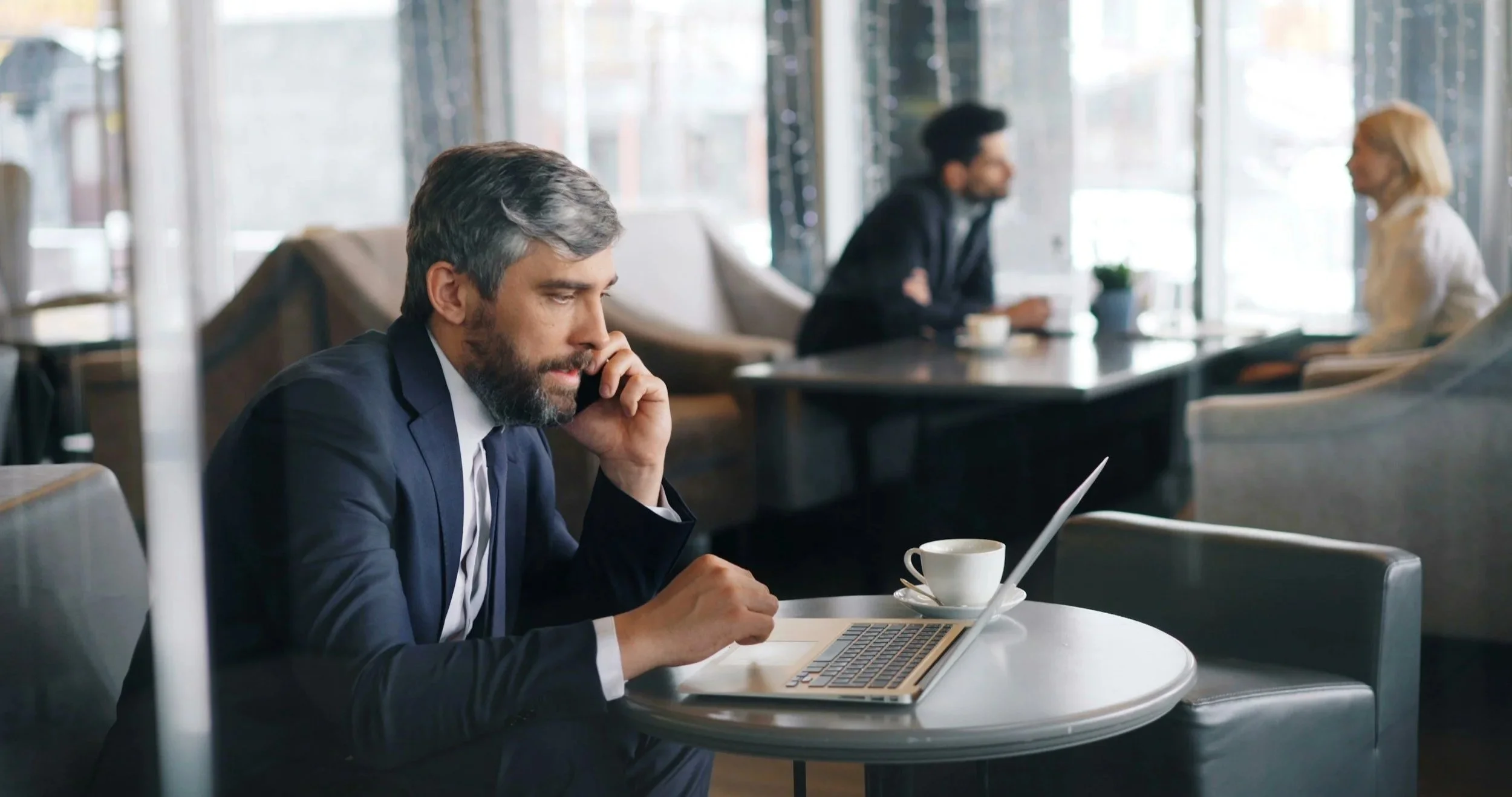 A man in a suit in a virtual meeting on a laptop at a cafe, with a coffee cup on the table.