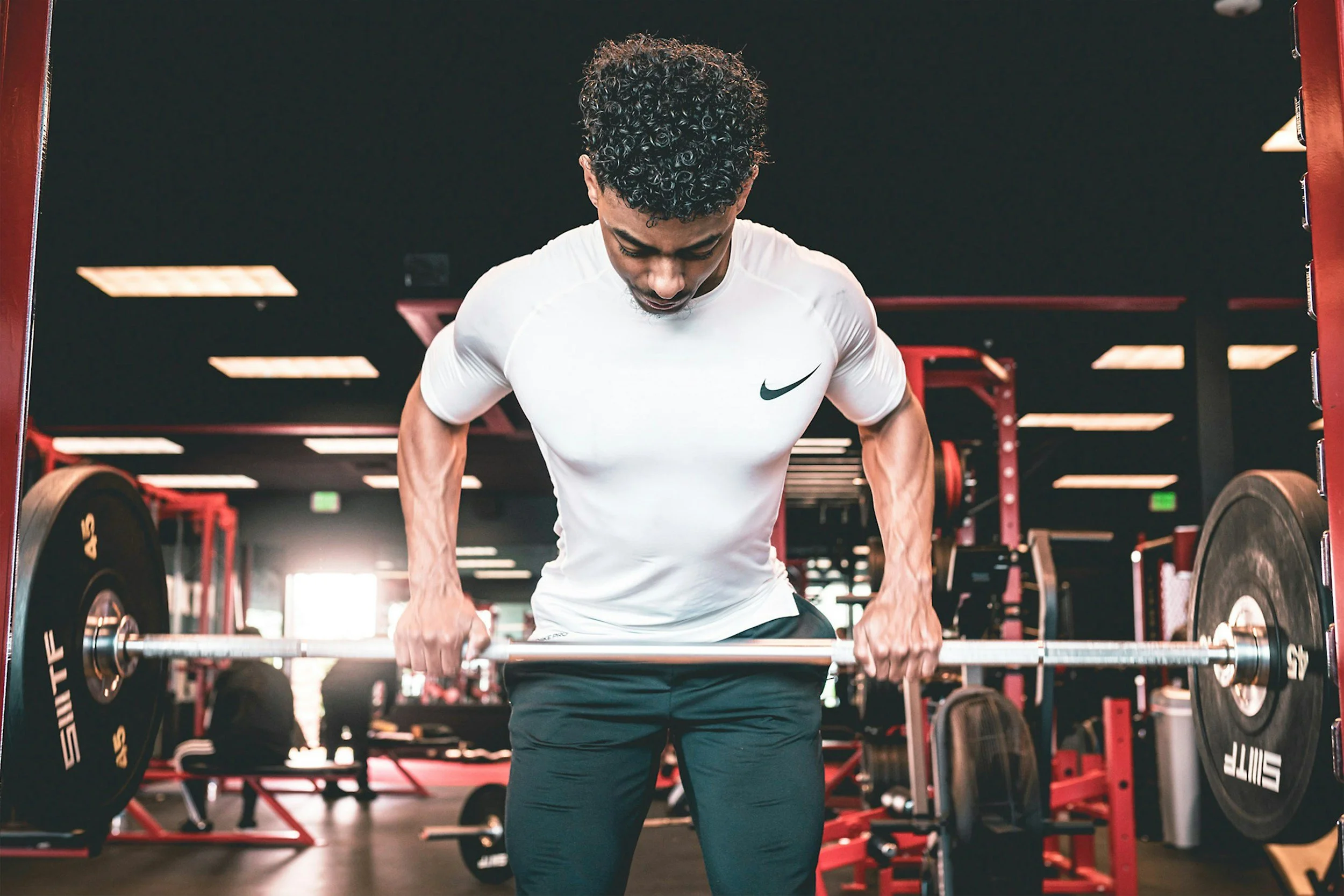 A young man with curly hair lifting a barbell in a gym.