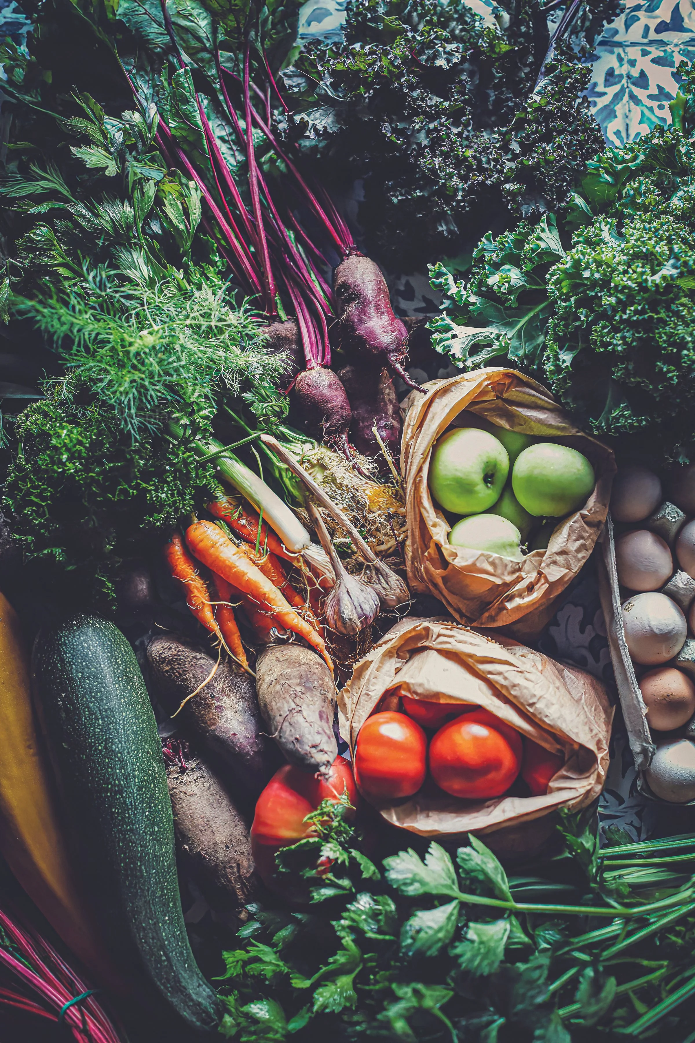 Un assortiment de légumes frais, y compris des betteraves, des carottes, des pommes, des tomates, des œufs, des courgettes, du chou frisé, des herbes et autres légumes variés, disposés sur une surface.