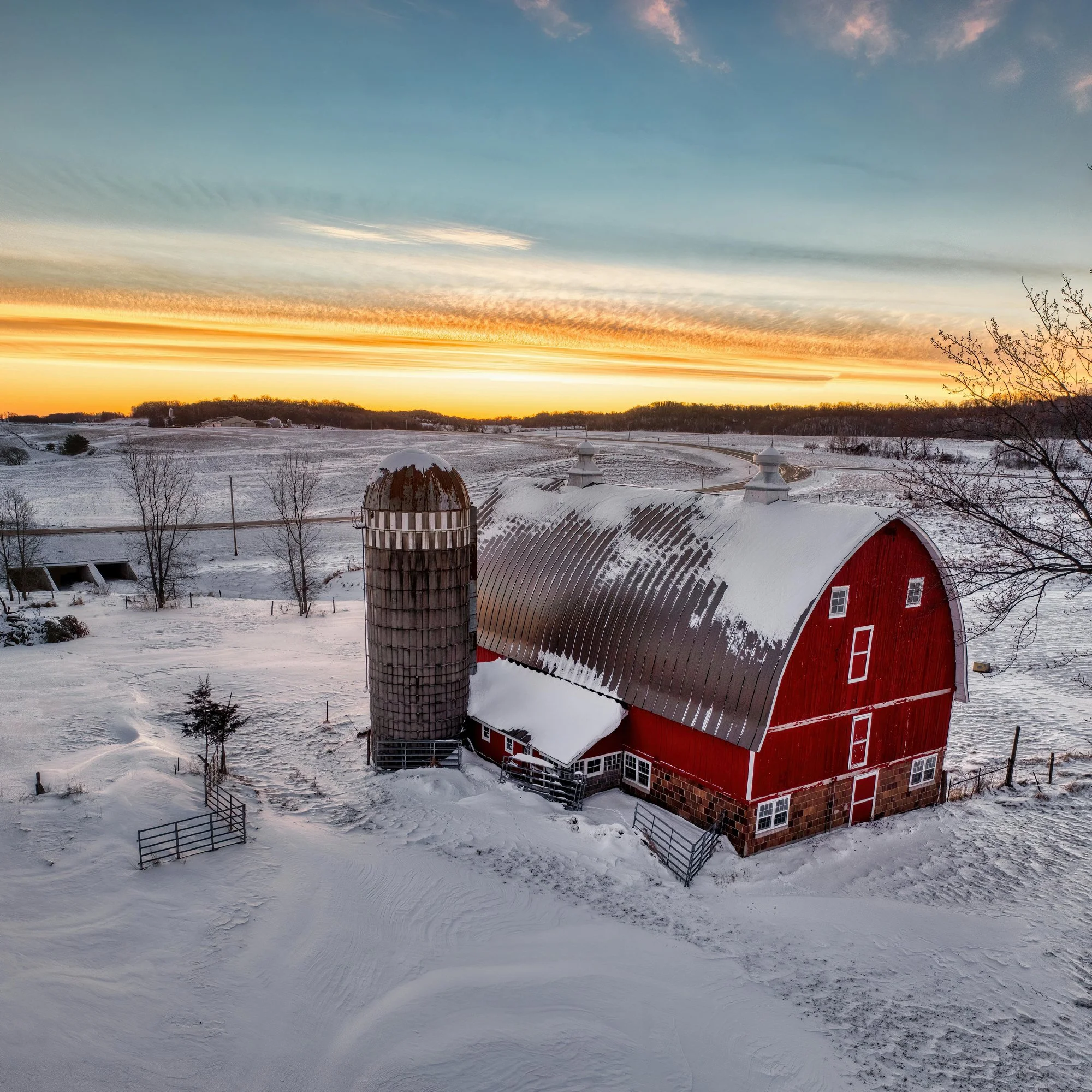 Vue d'une ferme avec un grand grange rouge et une grange en forme de silo dans un paysage enneigé au coucher du soleil.