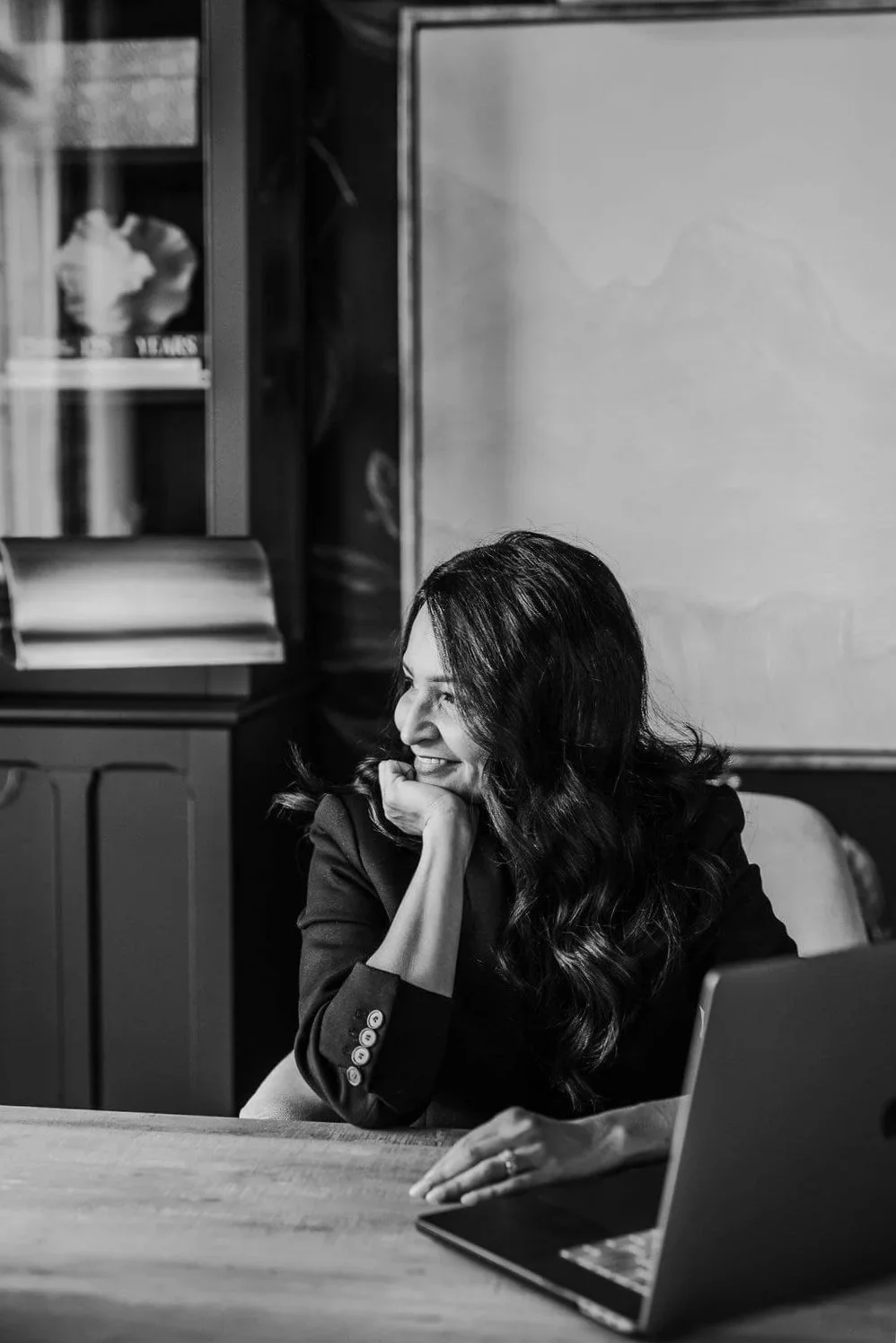 Black and white photo of Jasmine Shah, founder of The Velvet Finch, smiling and resting her chin on her hand at a desk with a laptop