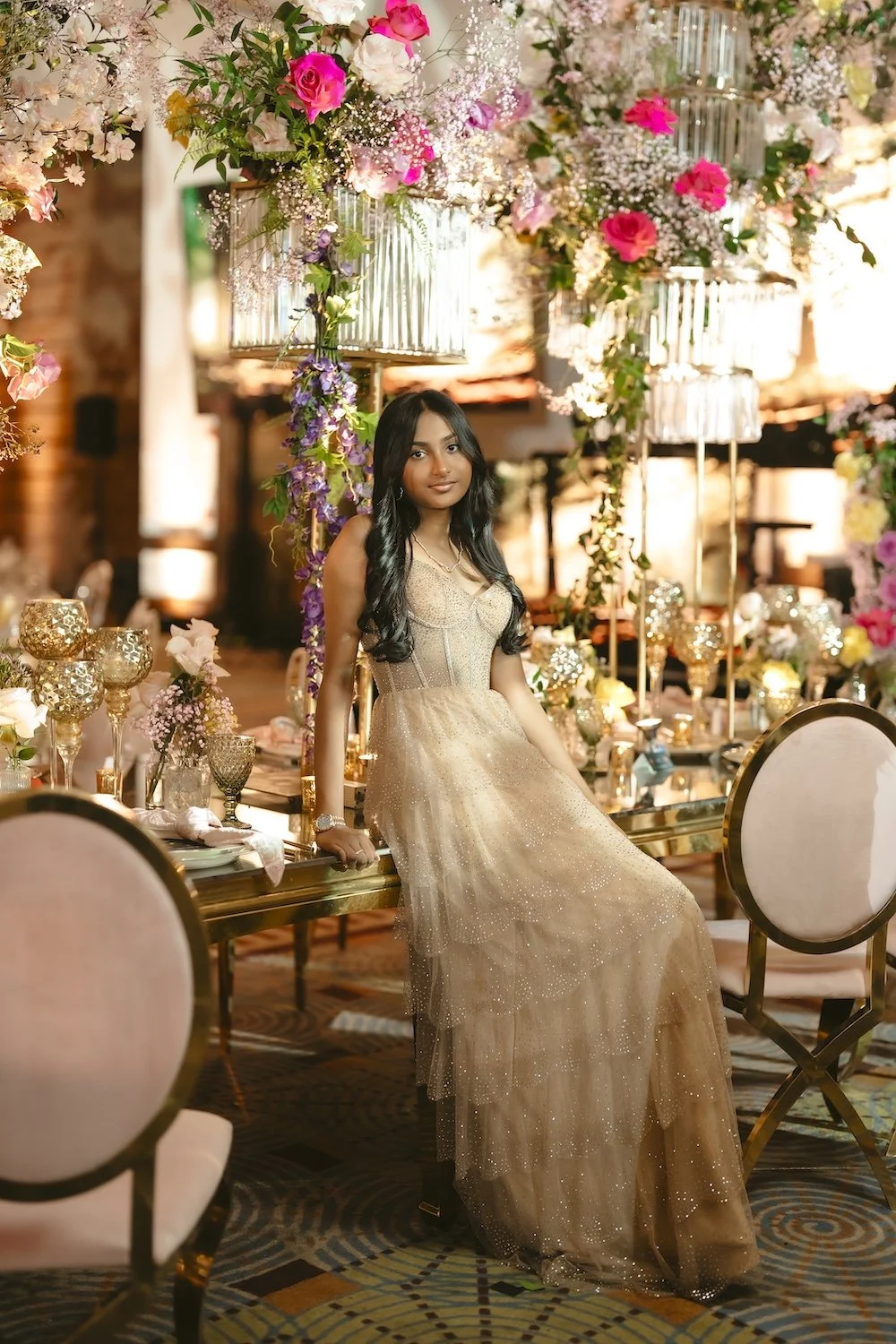 Young woman in a gold sparkle gown posing at a luxury event reception table surrounded by tall crystal chandeliers and cascading pink and purple floral arrangements