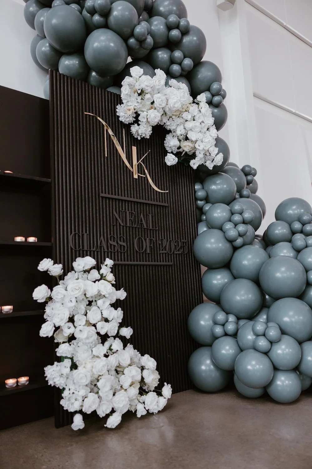Sage green and white balloon arch with white floral accents framing a custom NK monogram backdrop