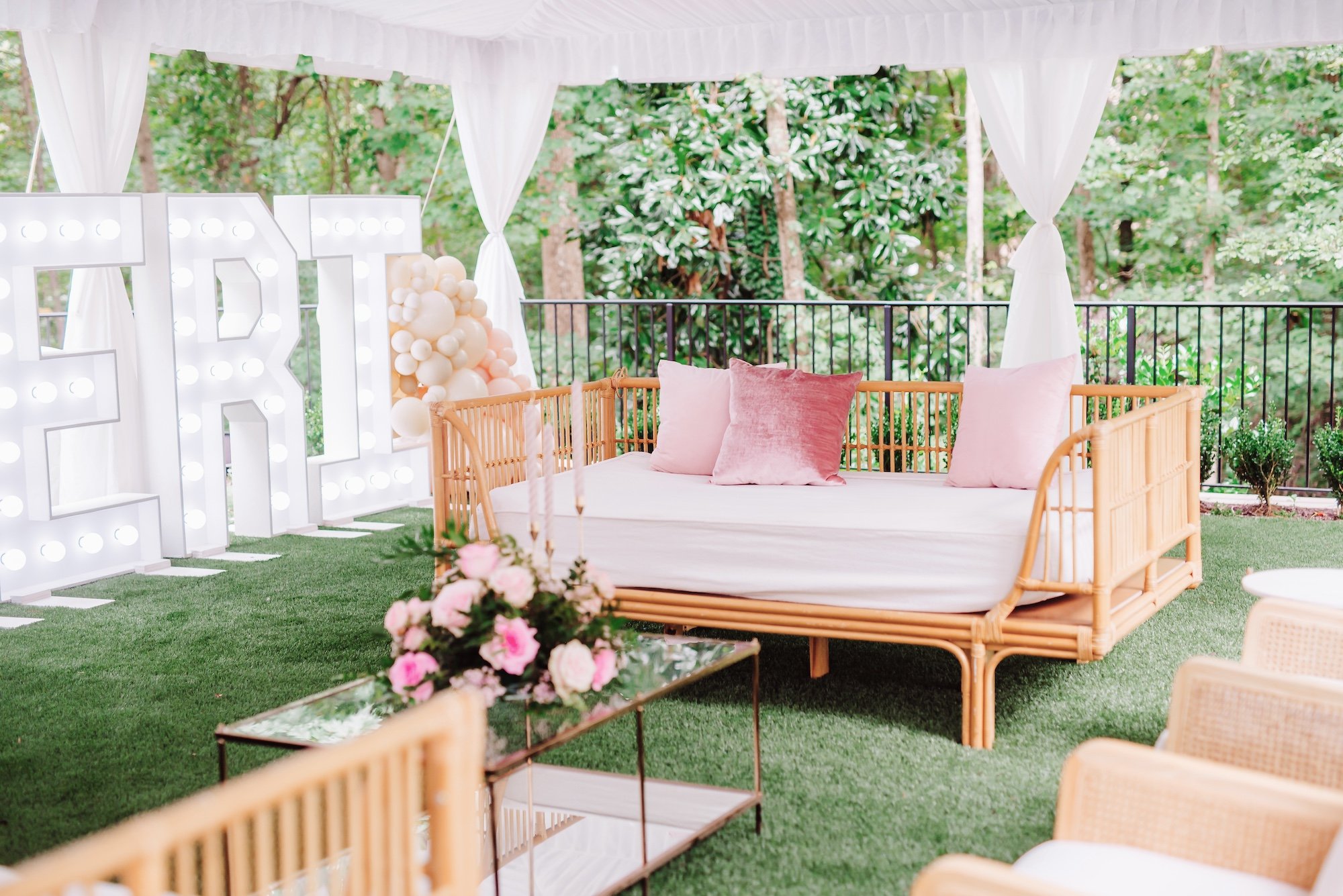 Outdoor garden party lounge area with a rattan daybed, pink velvet cushions, and pink rose floral arrangement on a mirrored coffee table, next to illuminated marquee letters and a cream balloon garland