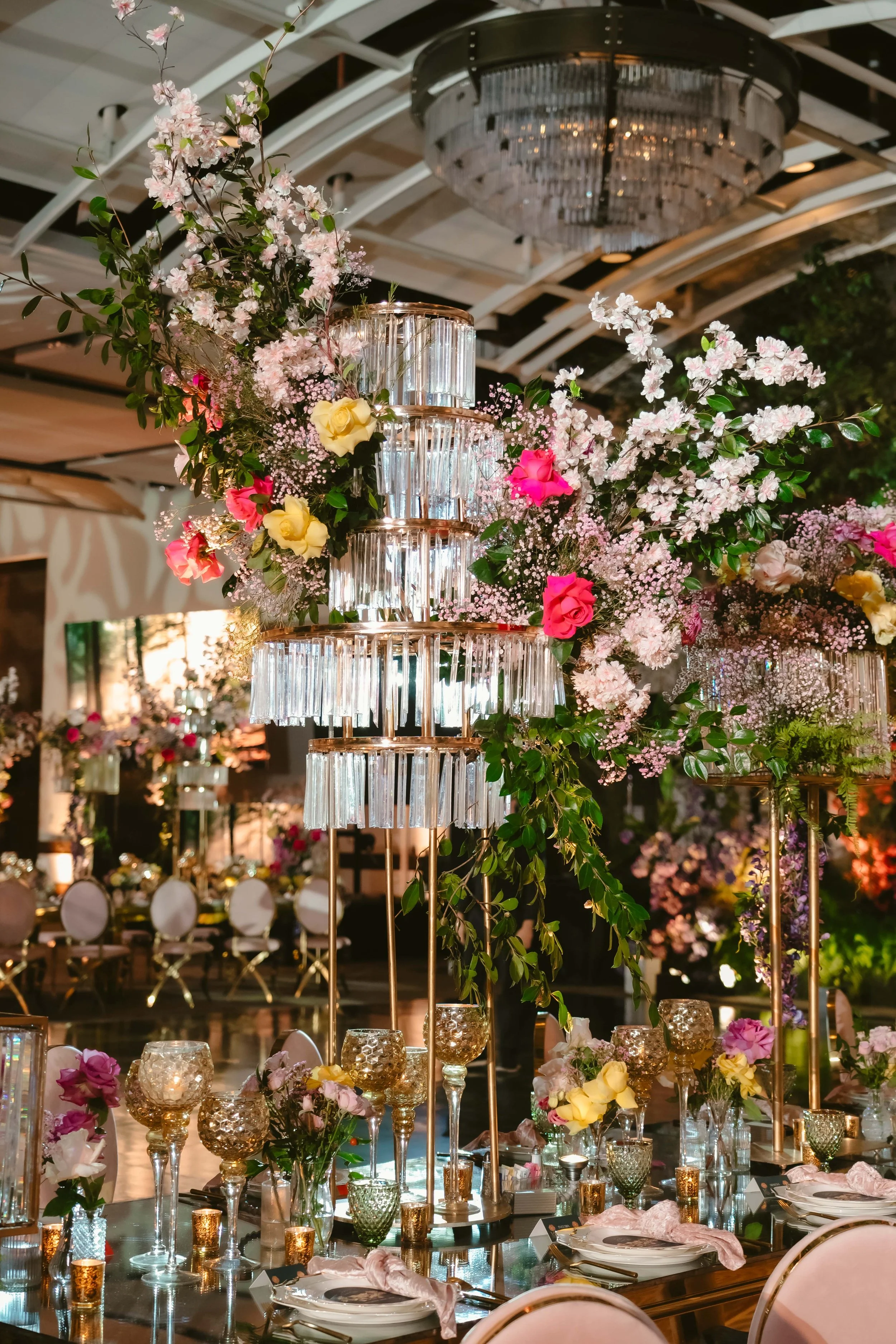 Tall tiered crystal chandelier centerpiece surrounded by lush pink and yellow floral arrangements at a luxury event