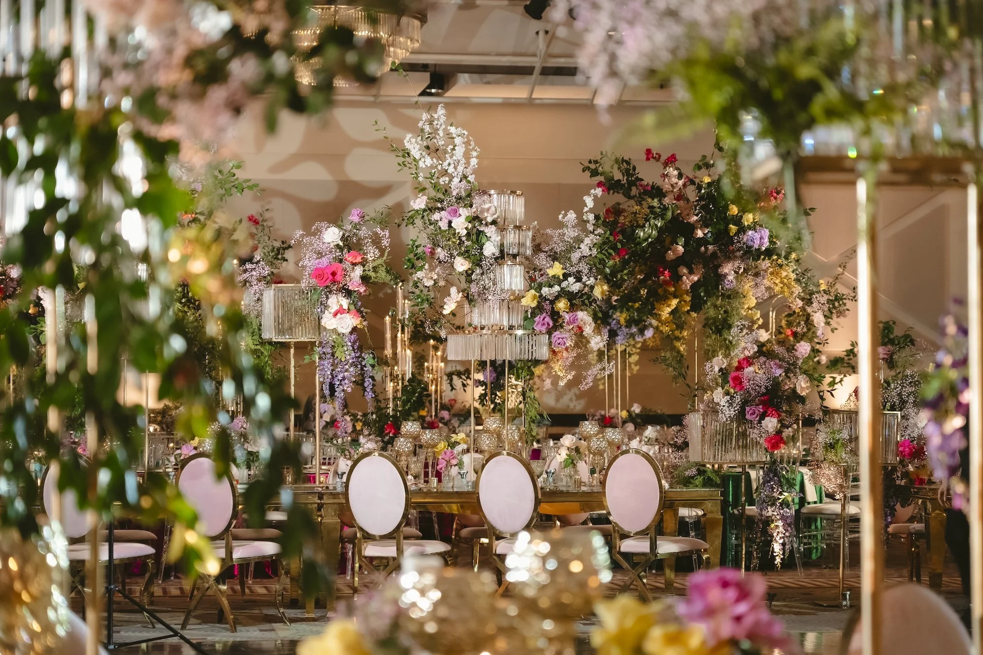 Luxurious wedding reception tablescape with towering crystal chandeliers, cascading floral arrangements in pink, purple, yellow, and red, and gold-framed chairs in a grand ballroom