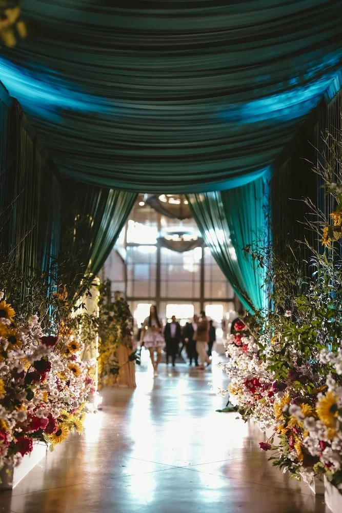 Dramatic event entrance corridor draped in teal fabric with sunflower and wildflower lined aisles and warm glowing light