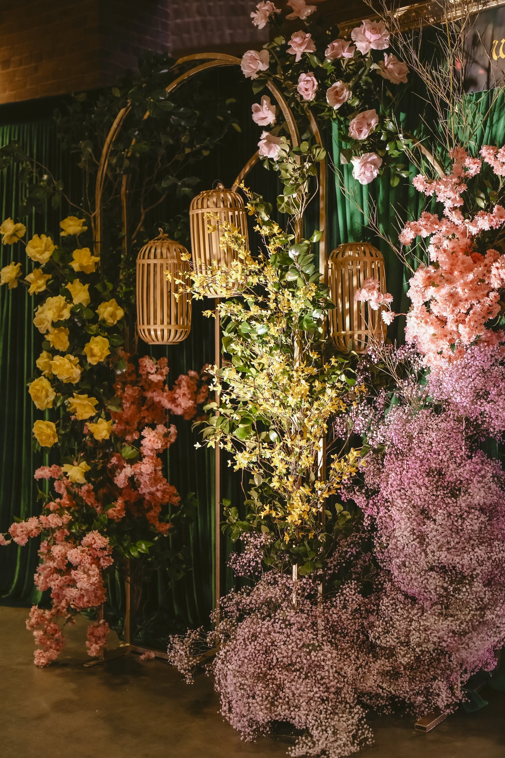 Lush garden-inspired event floral installation featuring yellow roses, pink blooms, baby's breath, and woven bamboo lanterns against a deep green draped backdrop