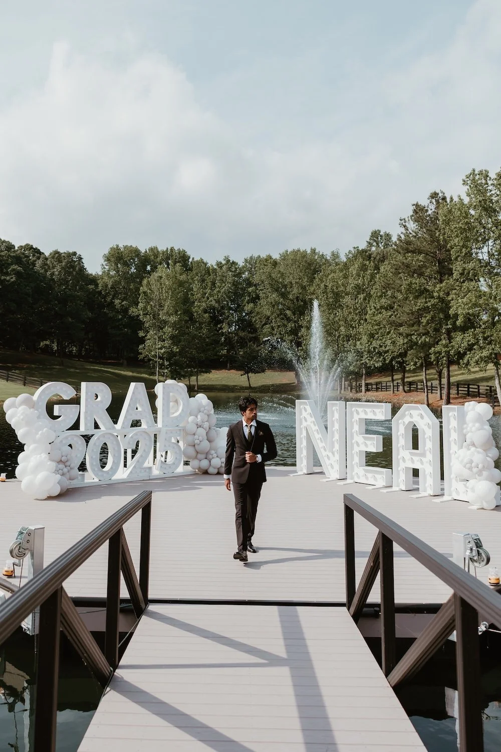 Graduate in a black suit walking across a lakeside dock at an outdoor graduation party with large marquee letters spelling GRAD 2025 and NEAL flanked by white balloon garlands and a water fountain