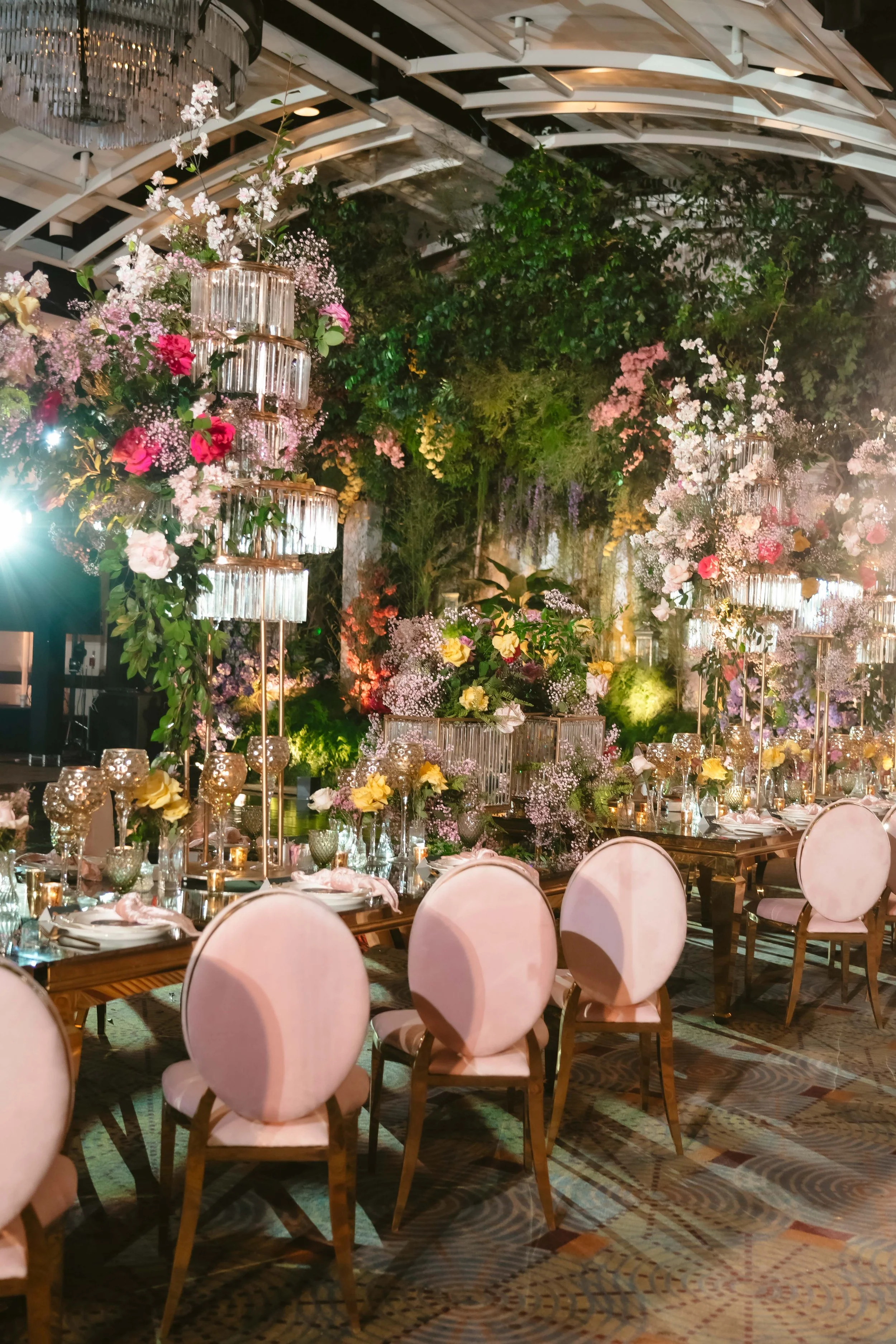 Lush garden-style wedding reception with tiered crystal centerpiece tower, colorful florals, and pink upholstered chairs at a candlelit dining table
