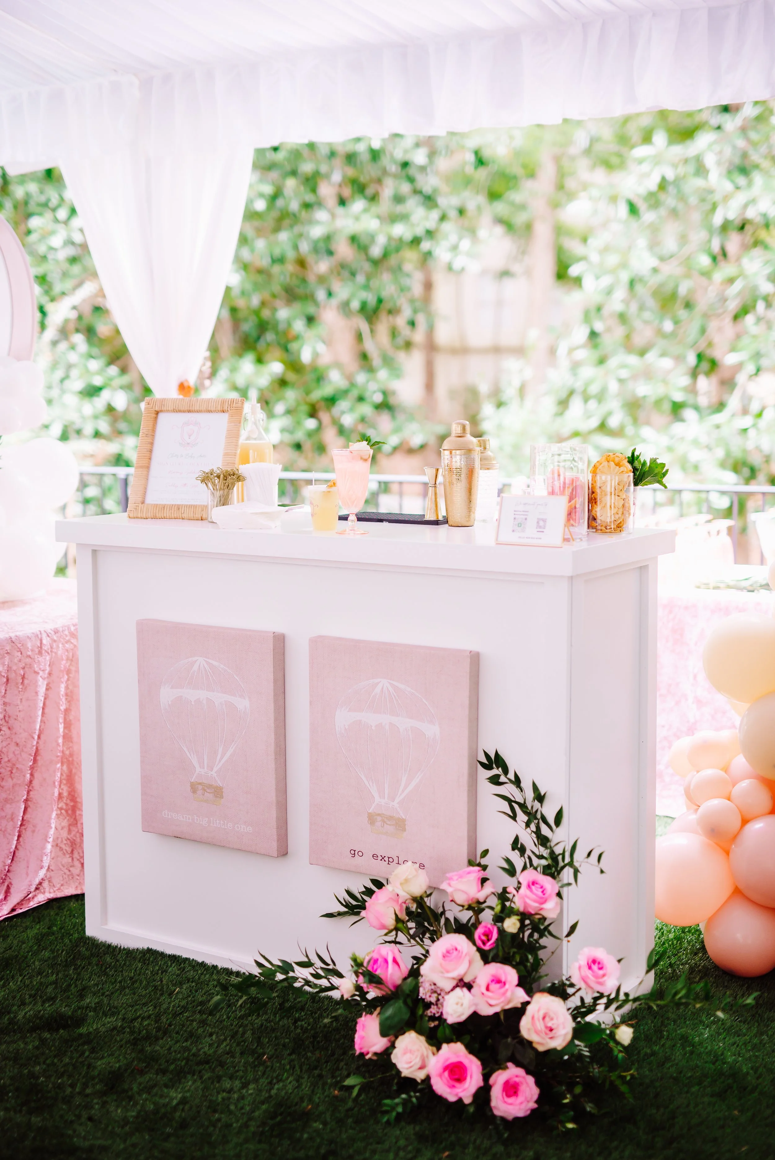 Styled outdoor event bar with white panels decorated with pink hot air balloon artwork, pink rose floral arrangement at the base, gold bar tools, and a peach balloon garland backdrop
