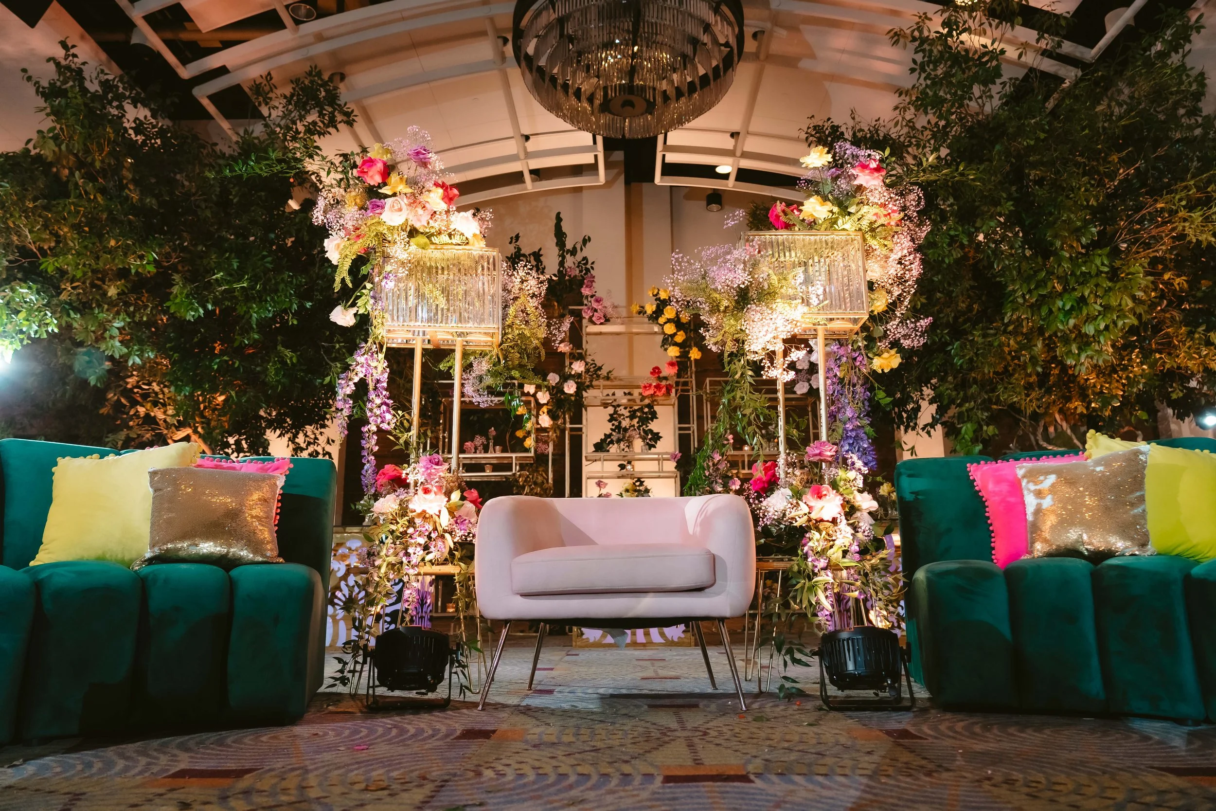 Dramatic event lounge with floor-to-ceiling floral installations in pink, purple, and yellow surrounding a blush loveseat and emerald velvet sofas under a crystal chandelier