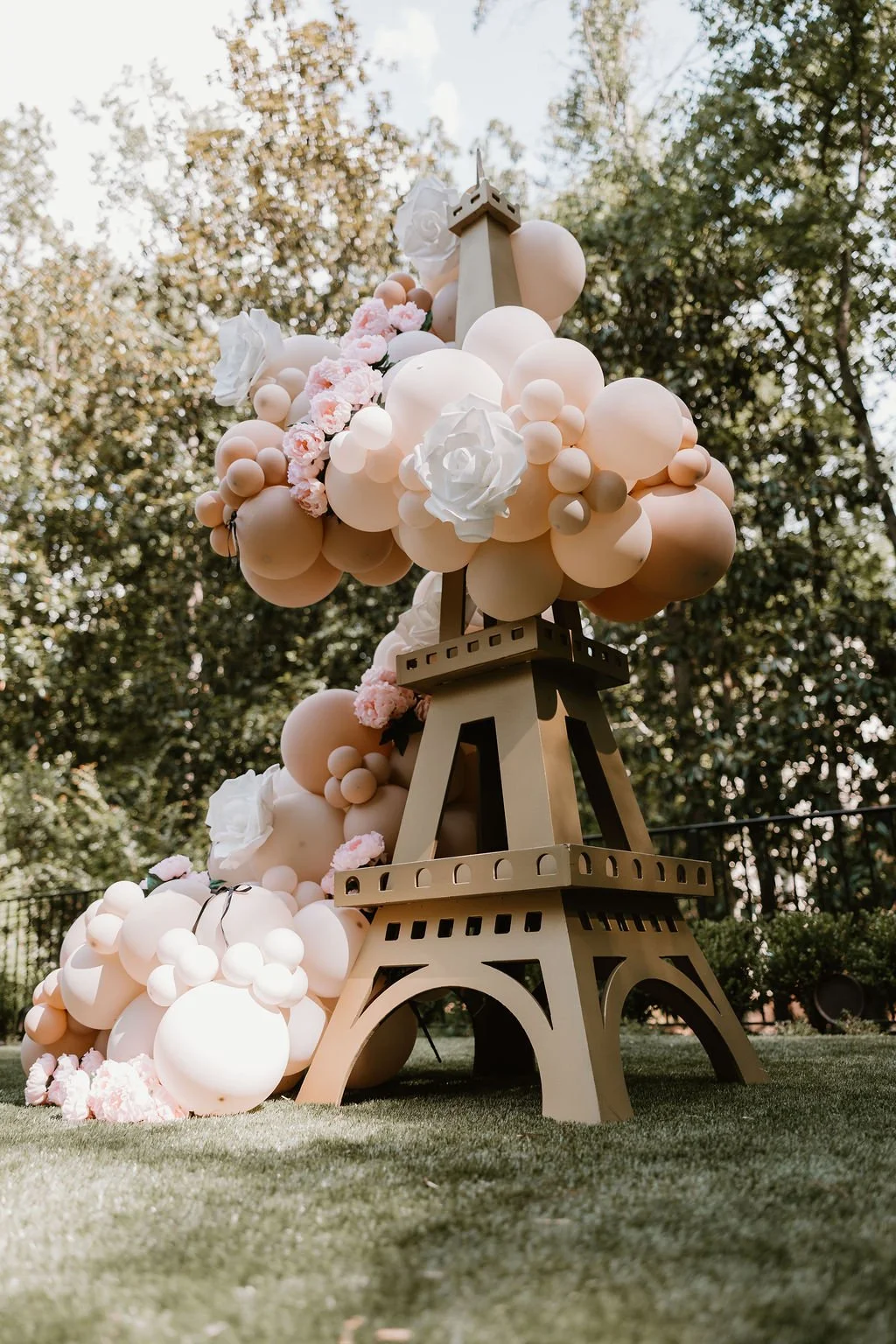 Outdoor event setup featuring a large gold Eiffel Tower prop covered in blush and cream balloon garland with oversized white paper roses