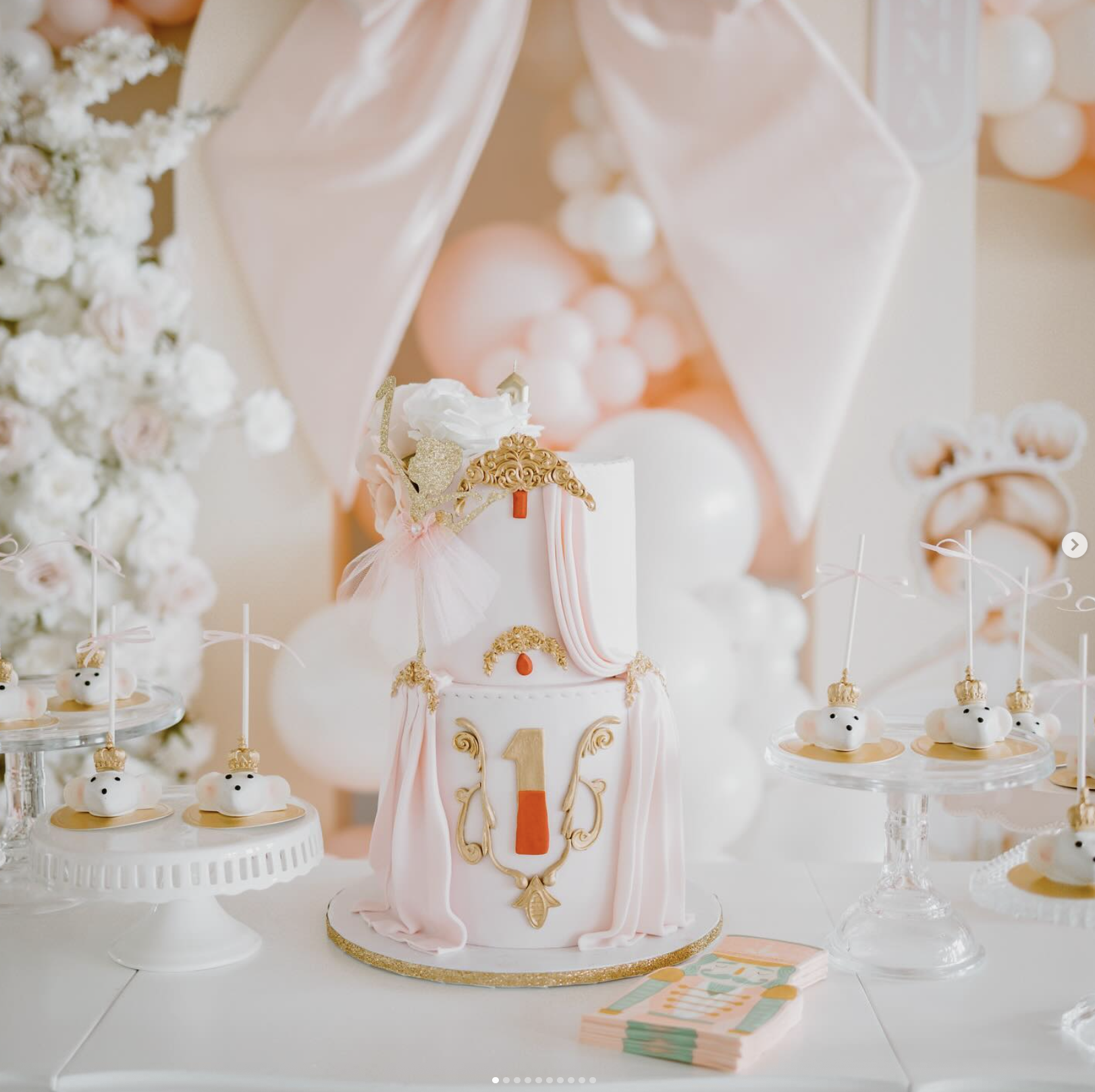 Ornate two-tier first birthday cake in blush pink and white with gold baroque details and draped fondant, surrounded by crown-topped cake pops on a white dessert table