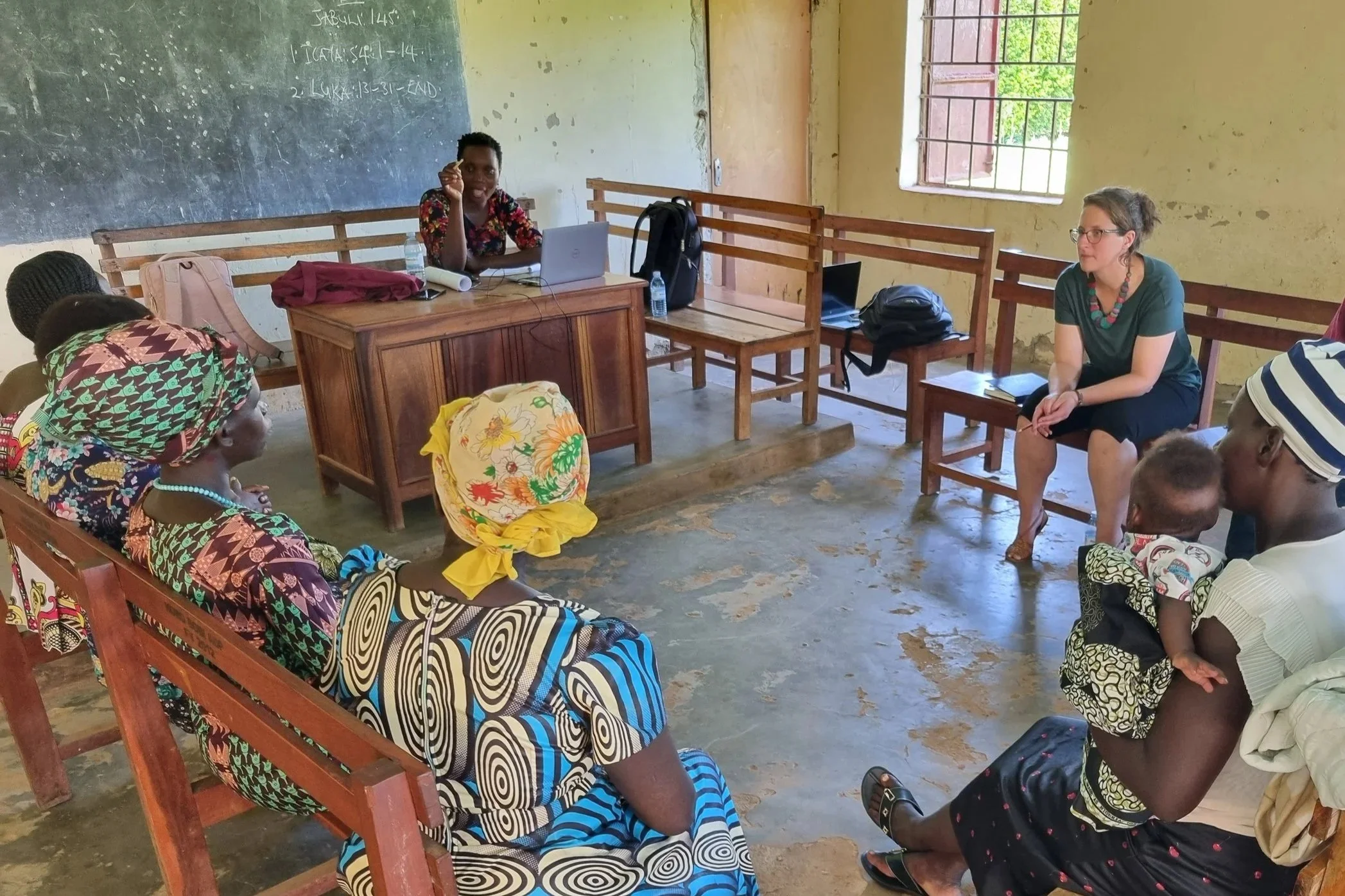 Photo of a workshop being held in an old classroom with a group of black women in colourful headscarves sitting on benches