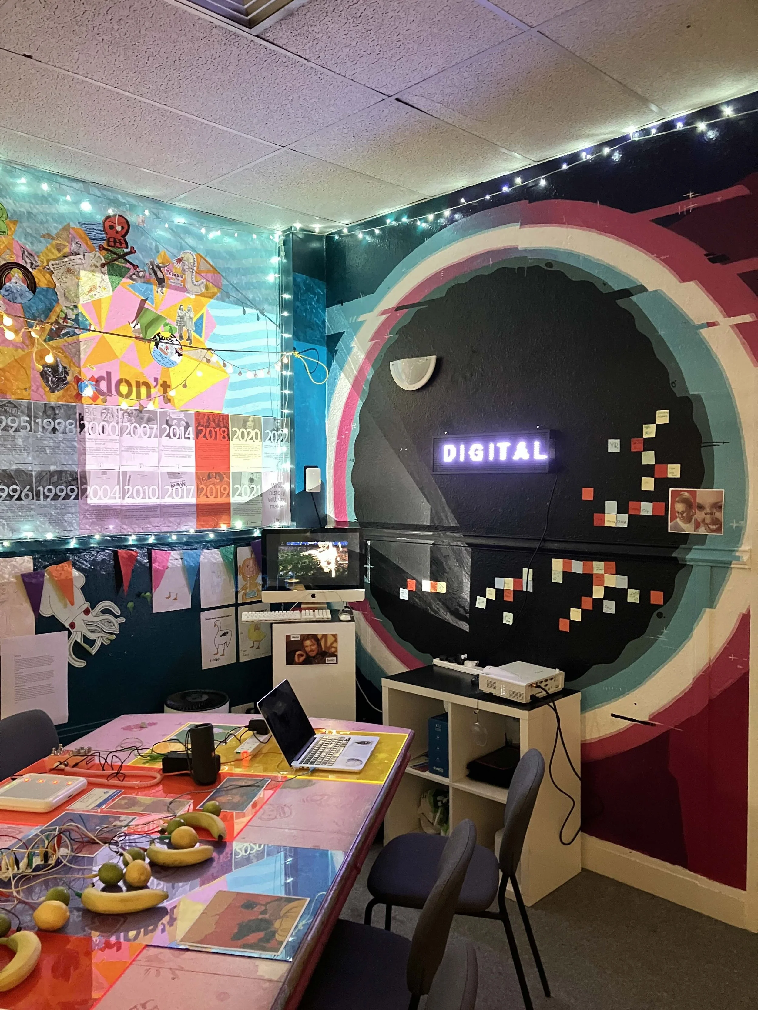 Photo of the corner of a room in a youth work setting with lots of colourful artwork on the walls. There is a laptop on a table in the foreground and a digital sign on the wall with the word 'DIGITAL'