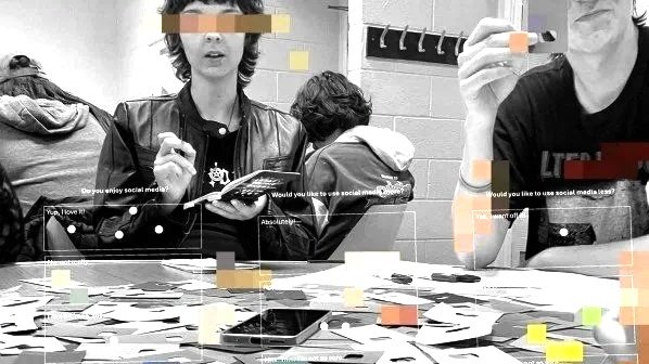 Black and white photo of a workshop with young people. One young person is facing the camera holding a reporter's notebook reading to take notes