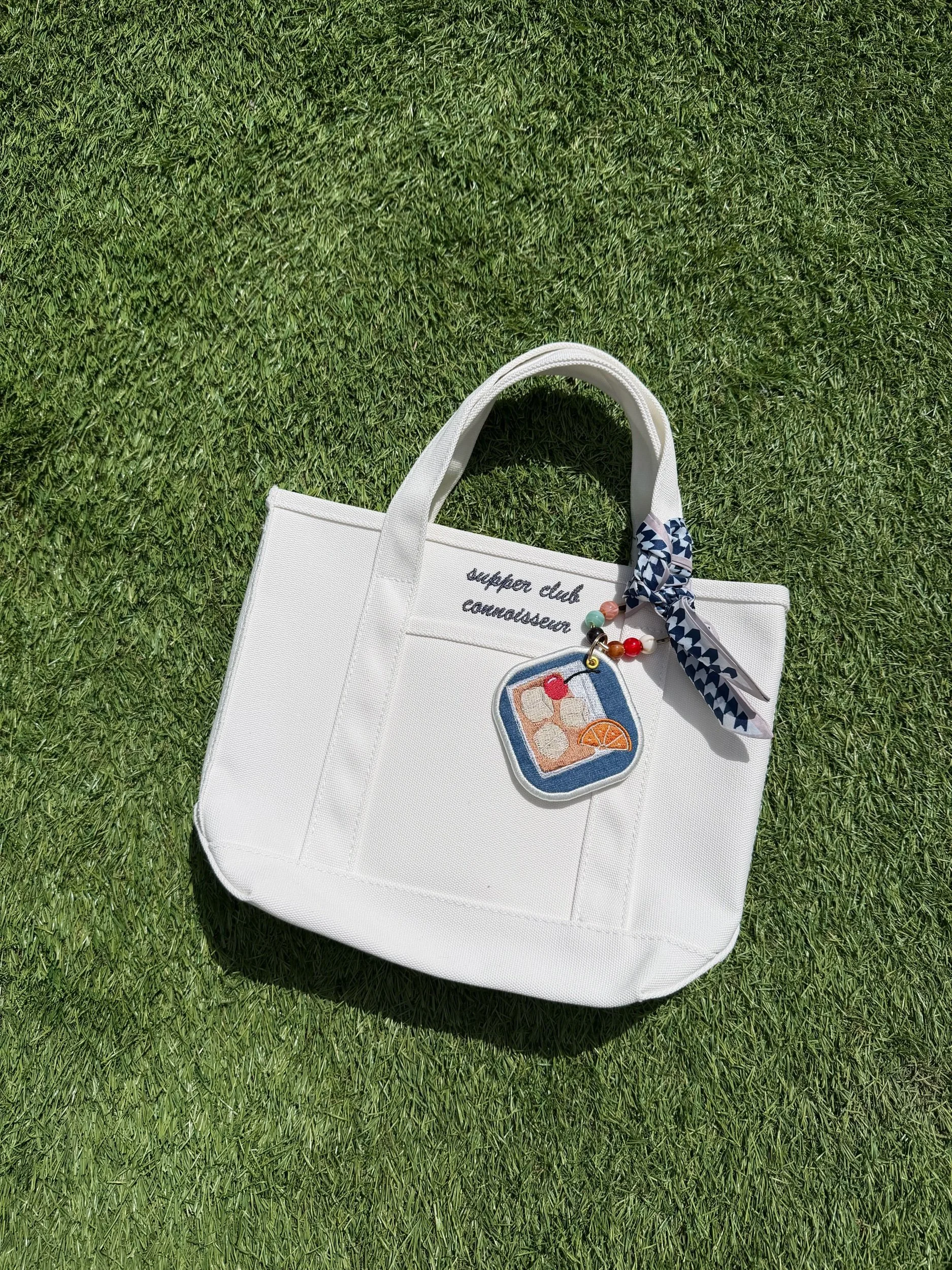 White canvas tote with supper club connoisseur on it and an old fashioned bag charm.