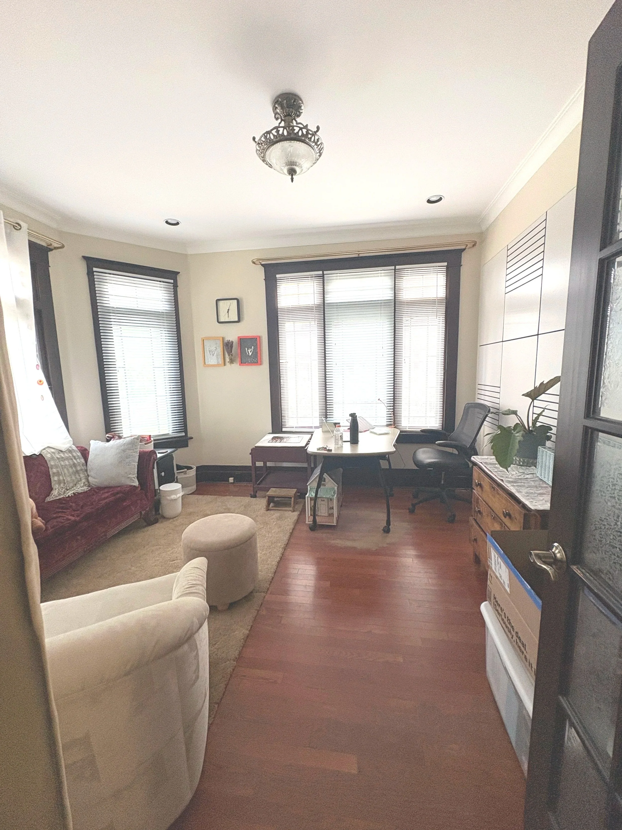 Living room with two large windows with black trim, a burgundy sofa with pillows, a beige armchair, a small round ottoman, a wooden dining table, a desk with a chair, plants, pictures, and a decorative ceiling light fixture.