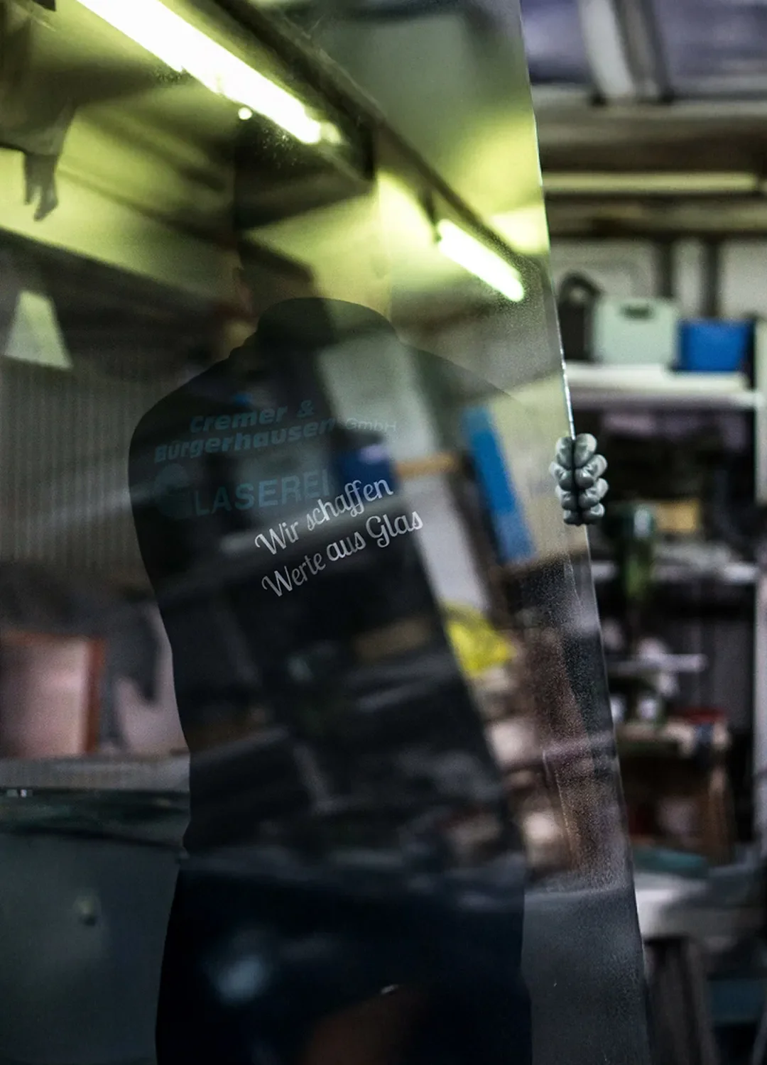 Mitarbeiter der Glaserei Cremer & Bürgerhausen hält eine große Glasscheibe in der Werkstatt; auf dem Glas spiegelt sich das Firmenlogo und der Slogan „Wir schaffen Werte aus Glas“.