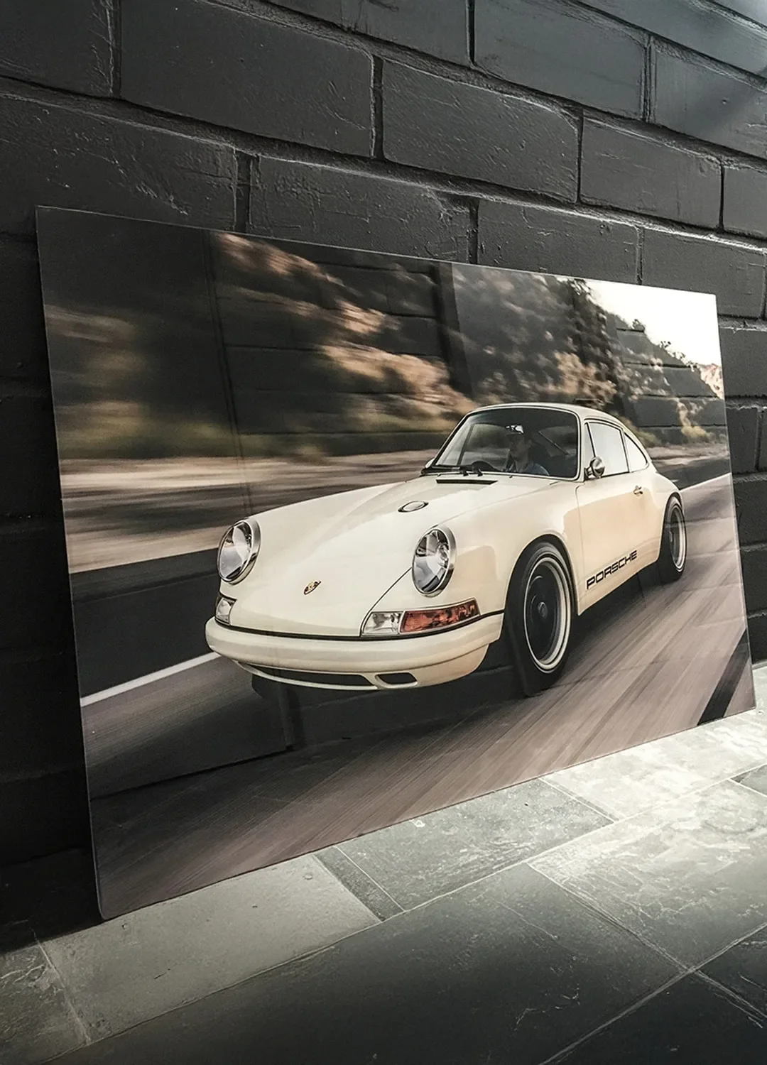 Beispiel für hochwertigen Glasdruck: Eine großformatige Glasscheibe mit fotorealistischem Aufdruck eines klassischen Porsche-Sportwagens, präsentiert vor einer schwarzen Ziegelwand.