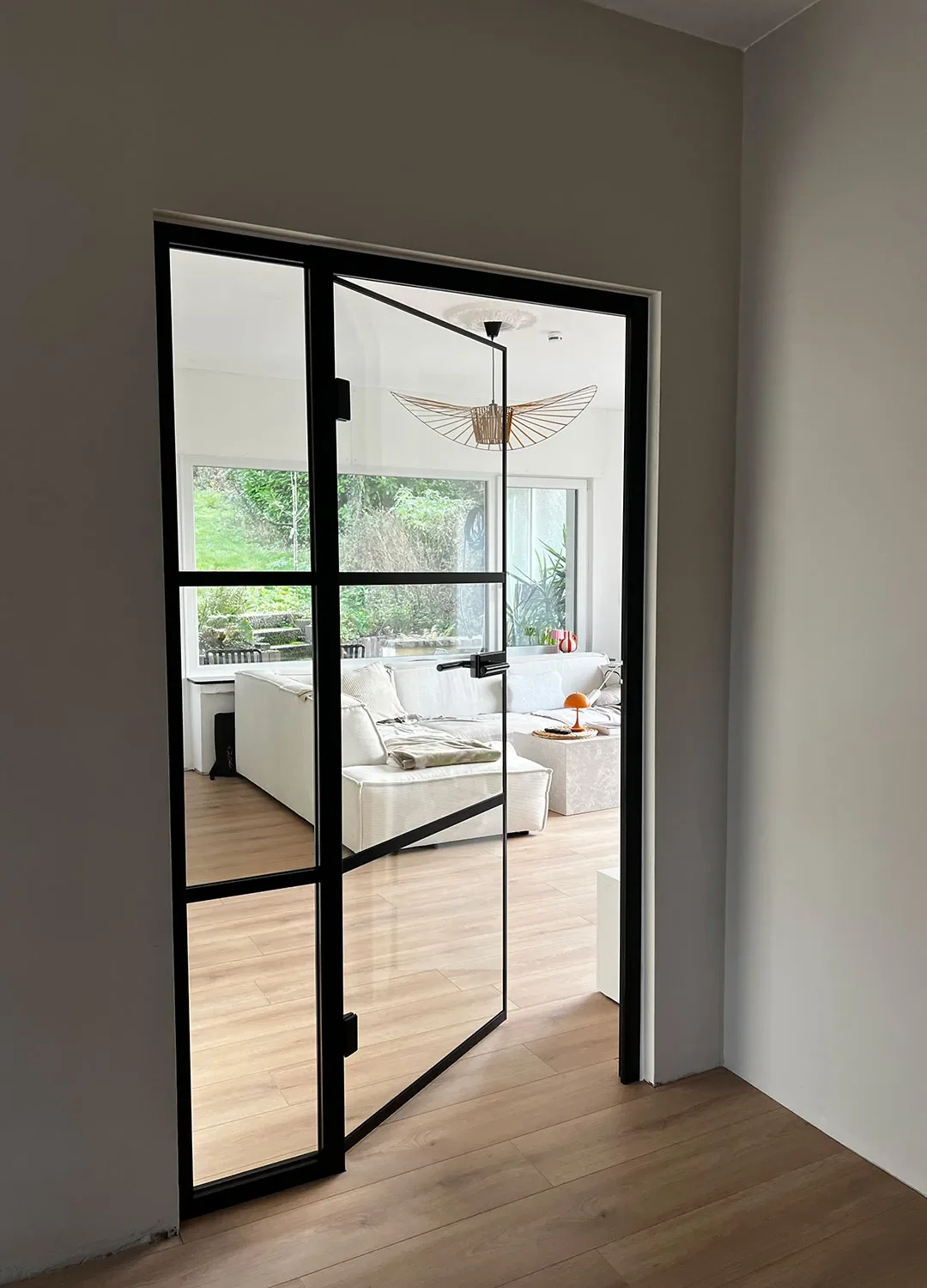 nnenansicht eines Flurs mit hellem Holzboden, der durch eine moderne, schwarze Stahl-Glas-Tür in ein lichtdurchflutetes Wohnzimmer führt. Die Tür steht offen und gibt den Blick frei auf ein weißes Ecksofa.