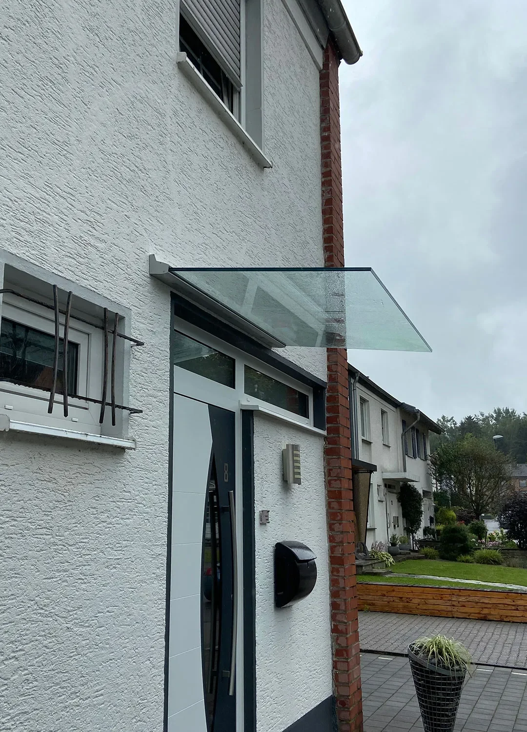 Modernes, rahmenloses Glasvordach über einer Haustür; die elegante Überdachung aus Sicherheitsglas wird durch eine dezente Wandschiene gehalten und schützt den Eingangsbereich vor Regen.