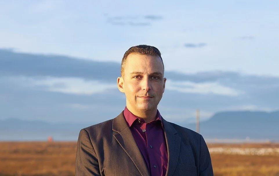 A man in a suit standing outdoors with a landscape and mountains in the background, during sunset or sunrise.