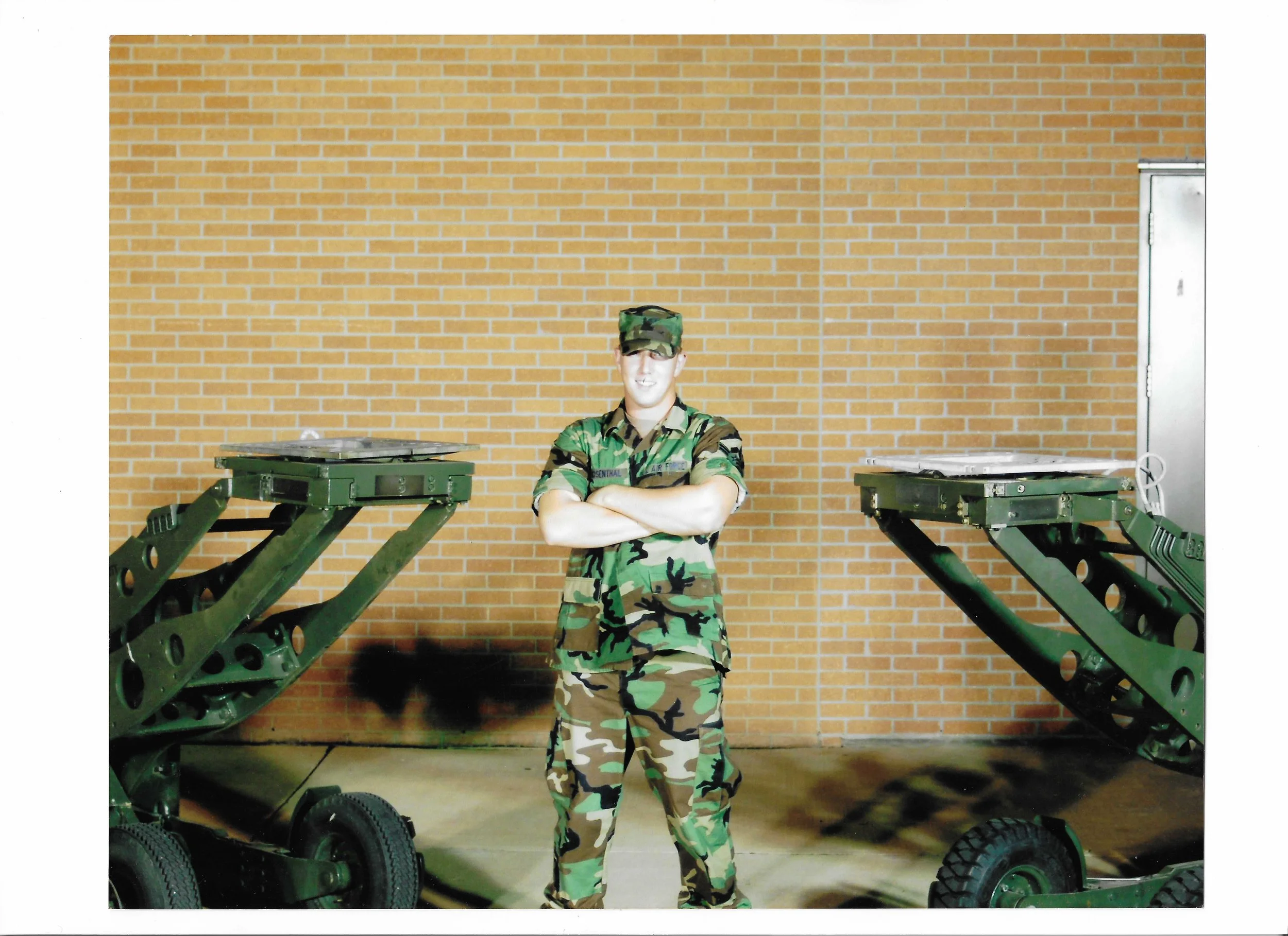 A person in a military uniform standing with arms crossed between two large green military equipment or carts against a brick wall.