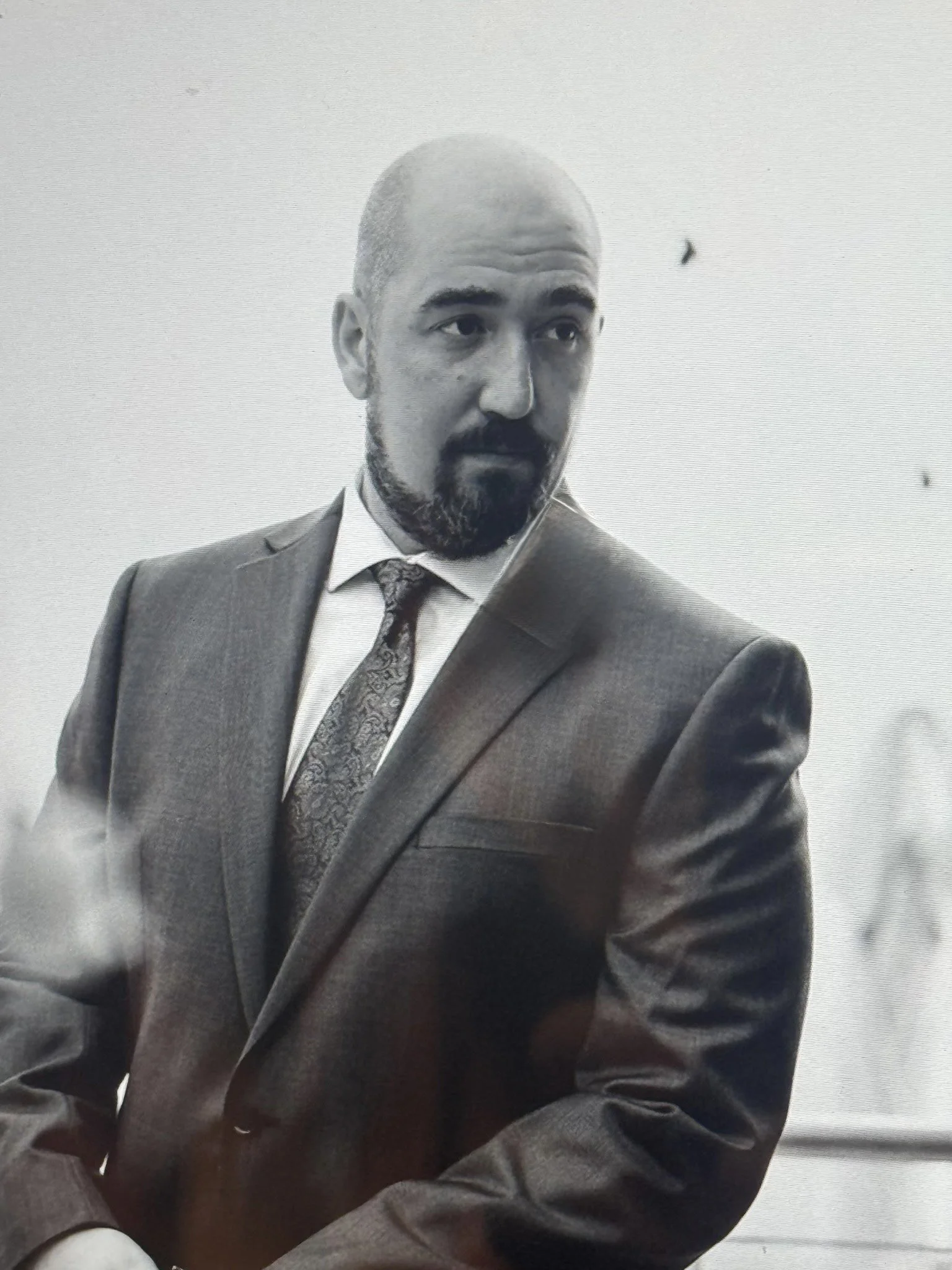 A man in a suit and tie with a bald head and beard, looking to his left, in a professional setting, black-and-white photograph.