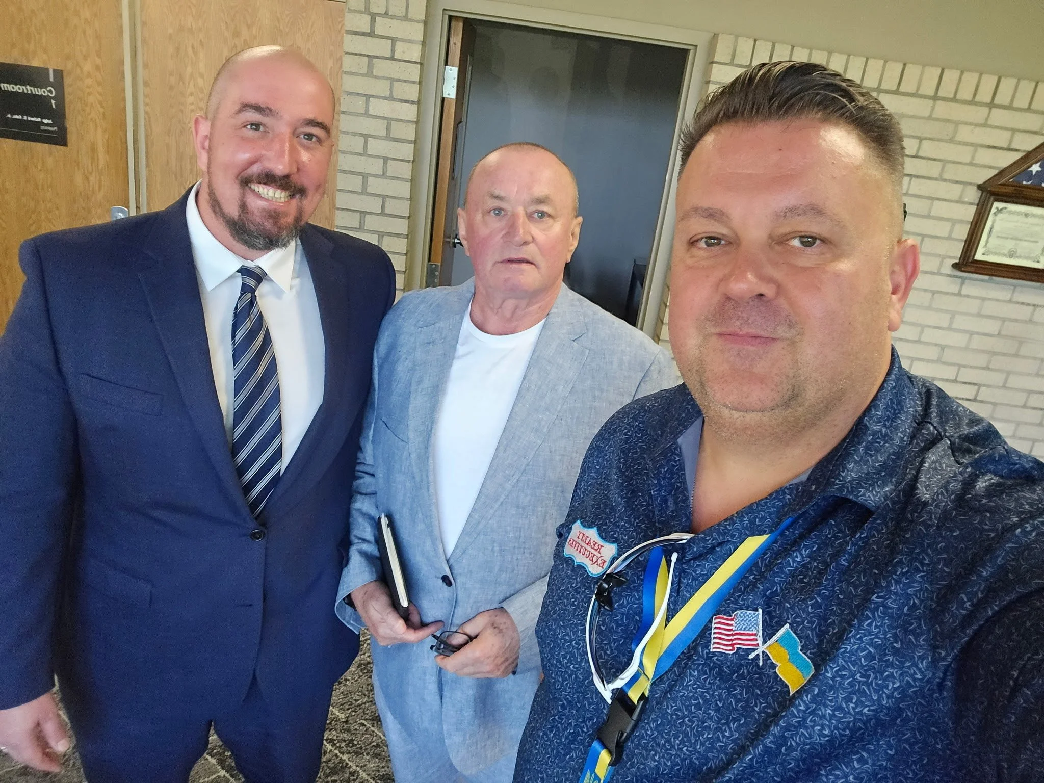 Three men in formal attire posing for a photo indoors, with a brick wall and certificates on the wall in the background.