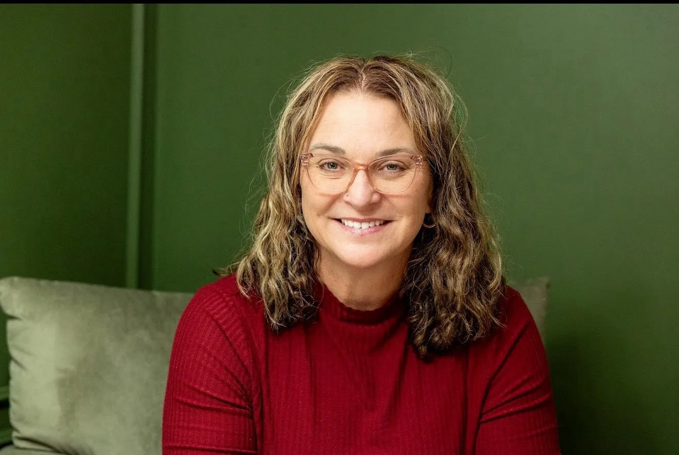 A woman with curly brown hair, glasses, wearing a red sweater, smiling, sitting on a green couch in front of a green wall.