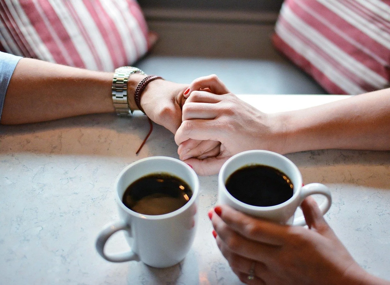Two people holding hands across a table with two cups of coffee, with a cozy background of striped cushions.