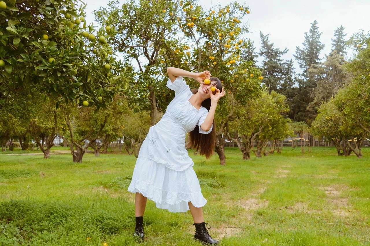 A woman in a white dress and black boots standing in an orchard, holding lemons near her face, with trees laden with lemons around her.