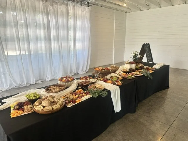 Buffet table with assorted snacks, fruits, and desserts, decorated with greenery, set up in a white-walled room with sheer drapes.