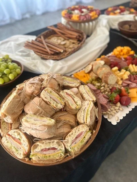 A platter of mini sandwiches with ham, cheese, and lettuce, surrounded by an assortment of fresh fruits, cheeses, and a charcuterie spread at a buffet table