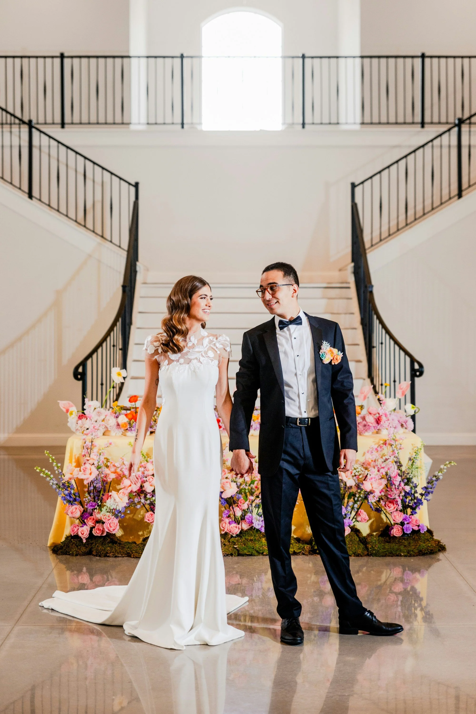 A bride and groom holding hands and looking at each other, standing in front of a floral wedding display inside a building with a staircase and large window in the background.