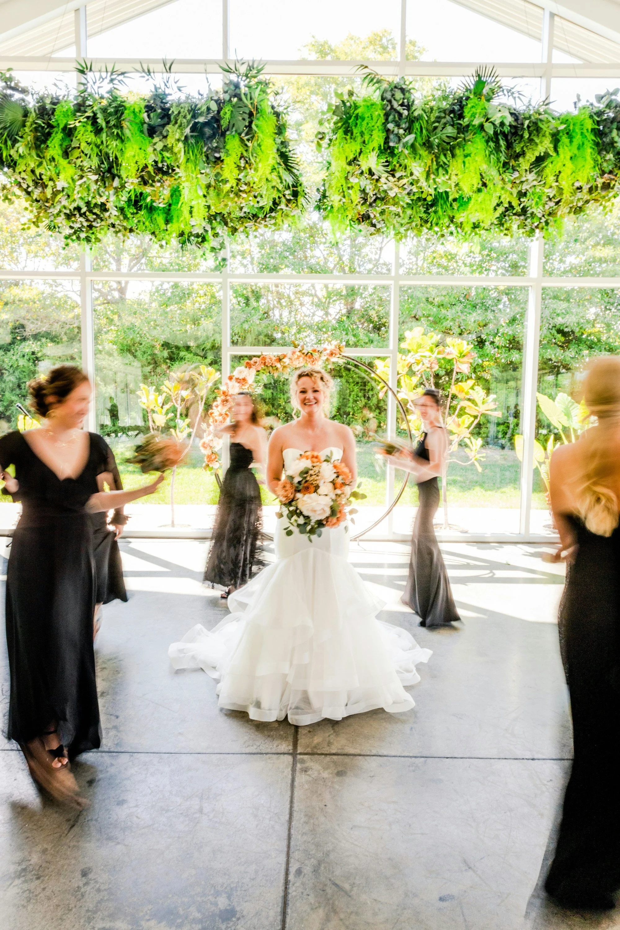 A bride with a bouquet of flowers in a white wedding dress, smiling, standing in front of a large window with greenery outside. Several women in black dresses surround her, some holding flowers, in a bright, modern wedding venue.