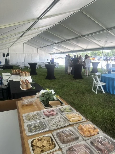 Inside a large event tent with food tables and guests mingling, serving dishes on the table and people gathered near high-top tables