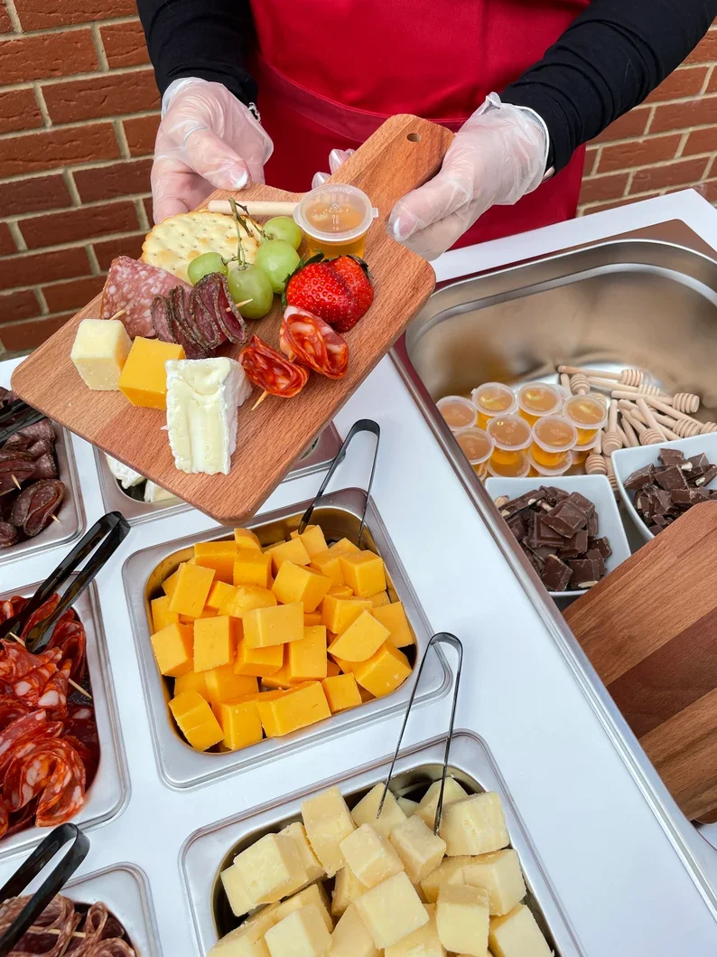Person wearing gloves and a red apron holding a cheese platter with grapes, strawberries, sliced meats, cheese, and a small container of honey, at a cheese and charcuterie station.